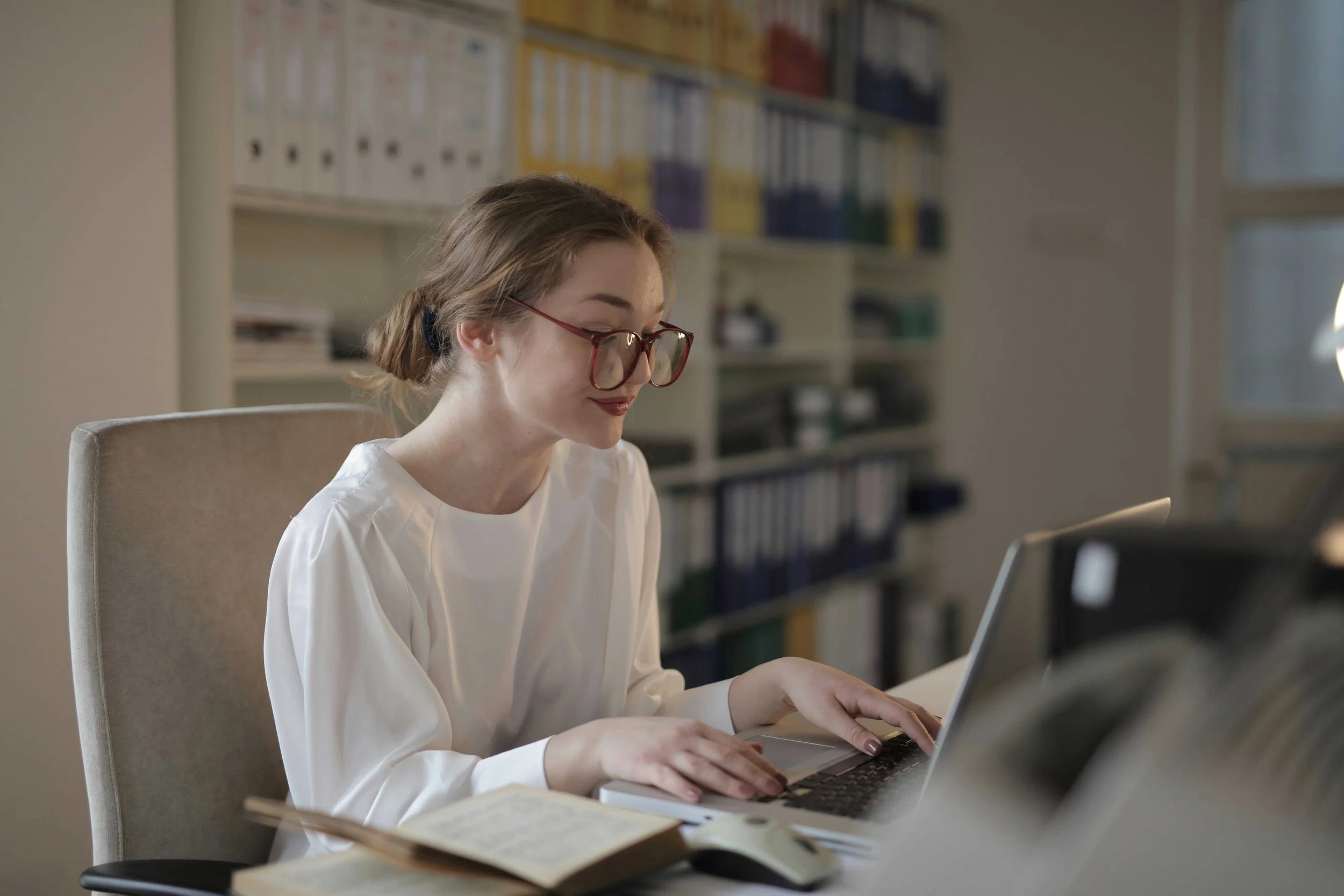 A young woman with glasses working on a laptop at a desk, with an open book and a computer mouse nearby, in an office or library setting.