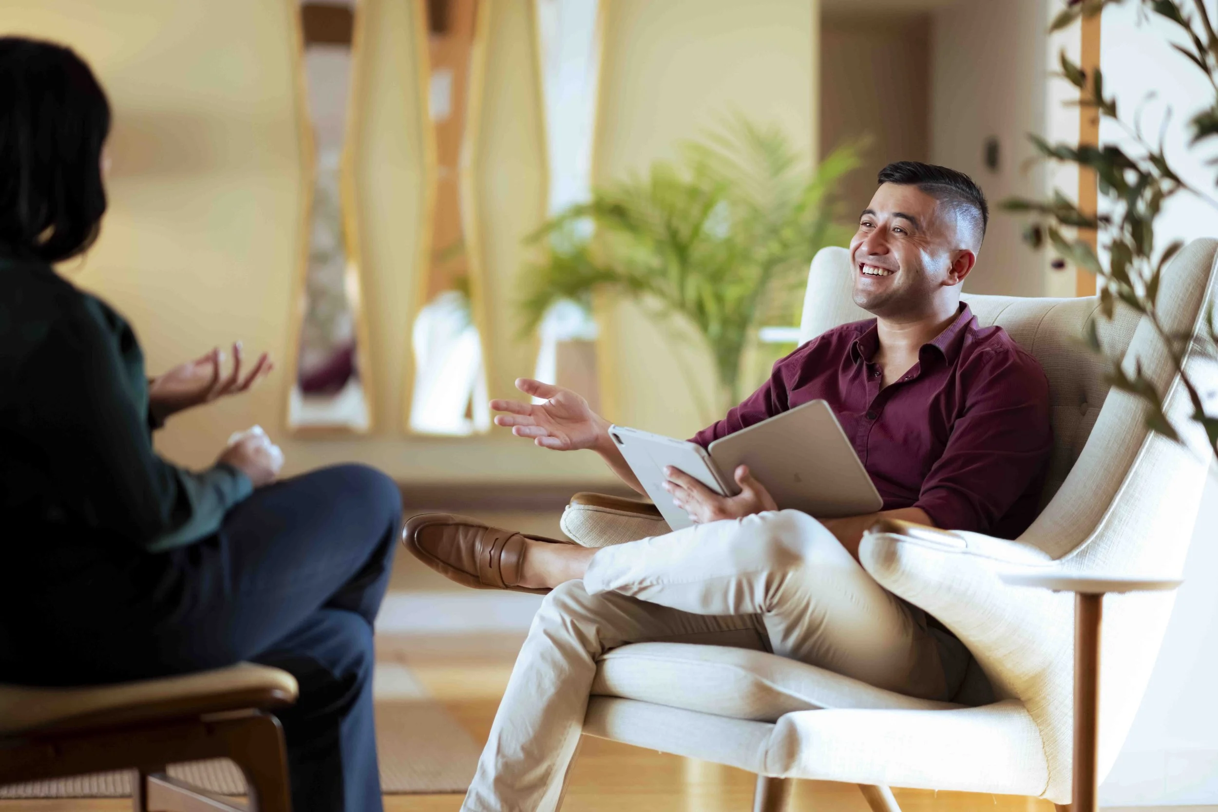 Daniel Mark Hipp, henderson therapist, having a therapy session in a cozy, well-lit room with plants, one person is seated on a chair with a notebook and the other is sitting on a sofa.