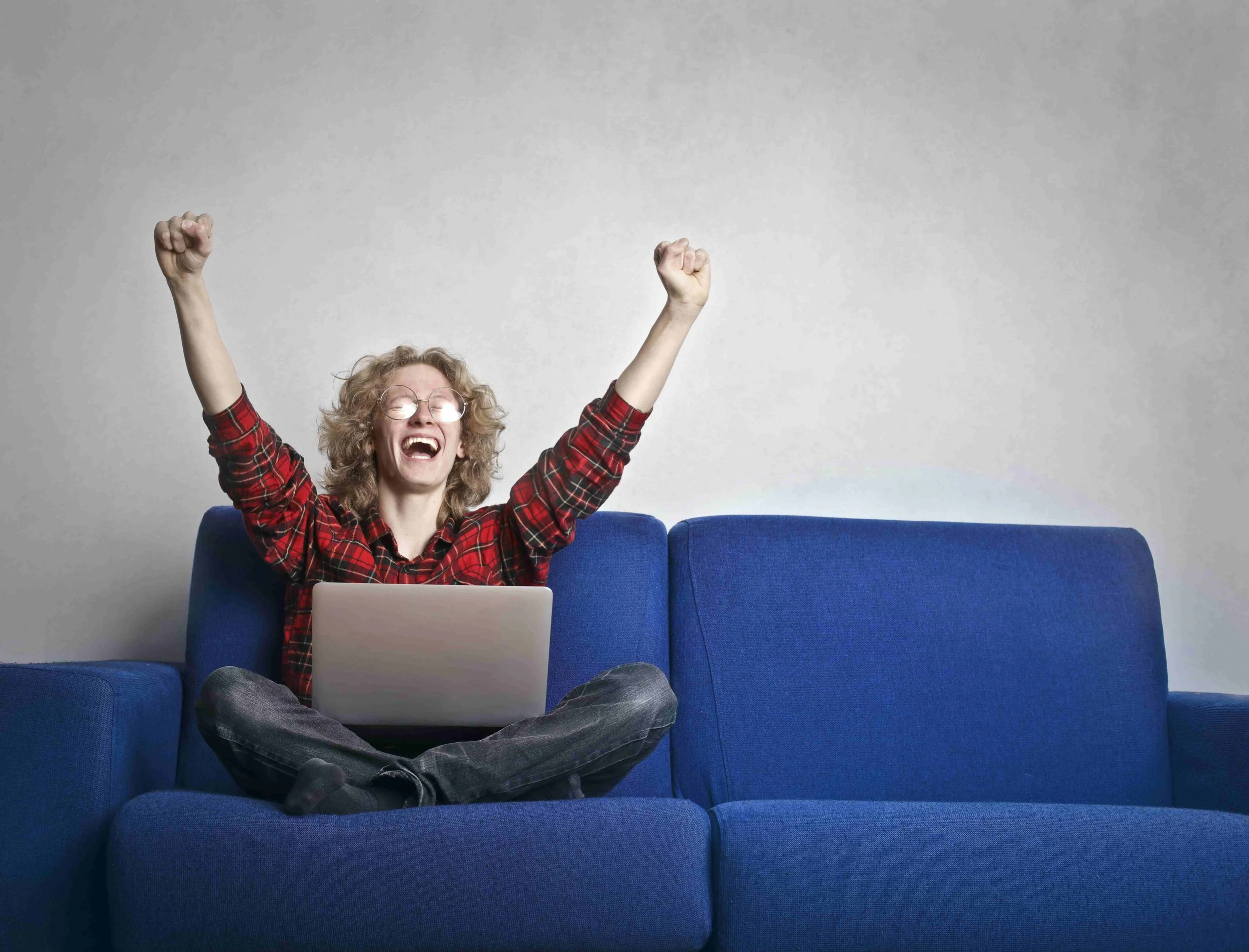 Person with glasses and curly hair sitting on a blue couch with a laptop, celebrating with arms raised and smiling.