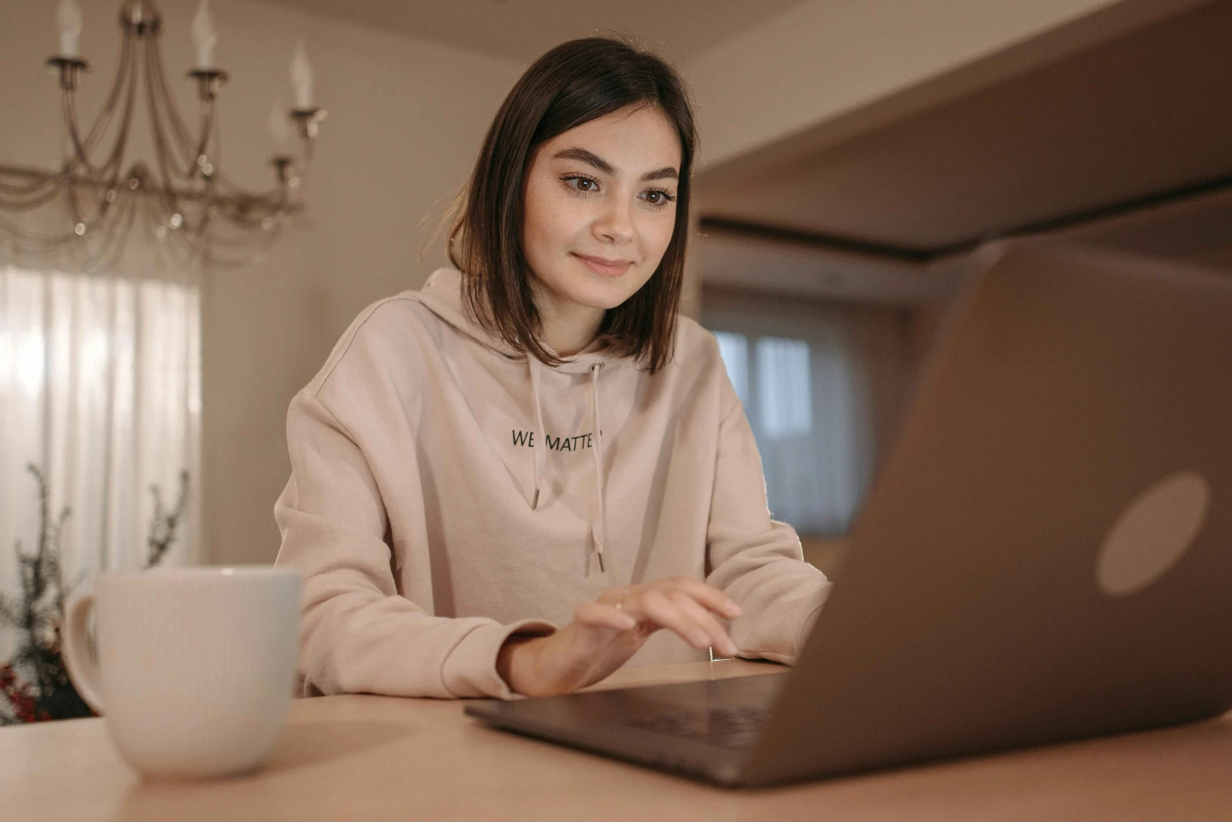 A young woman with shoulder-length brown hair attending an online therapy session. She is looking at her laptop, white mug in the foreground, chandelier hanging from ceiling in background, indoors with natural light coming through windows.