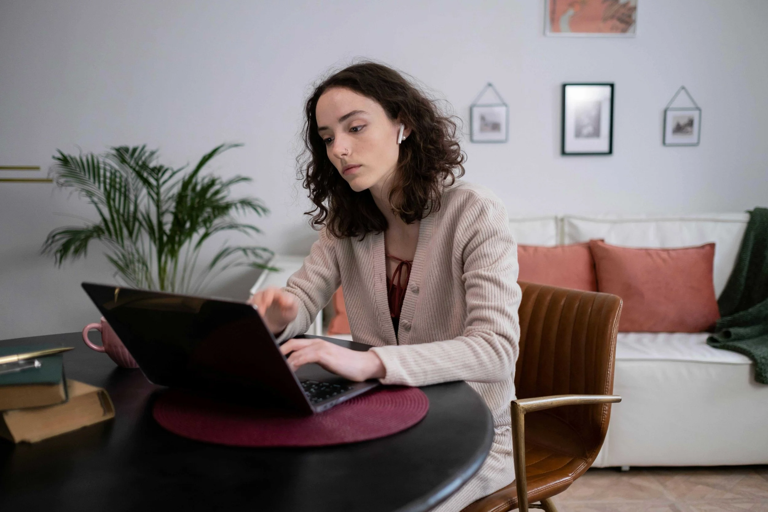 A young woman with curly hair working on a laptop at a dining table in a living room, with a plant and framed pictures on the wall behind her.