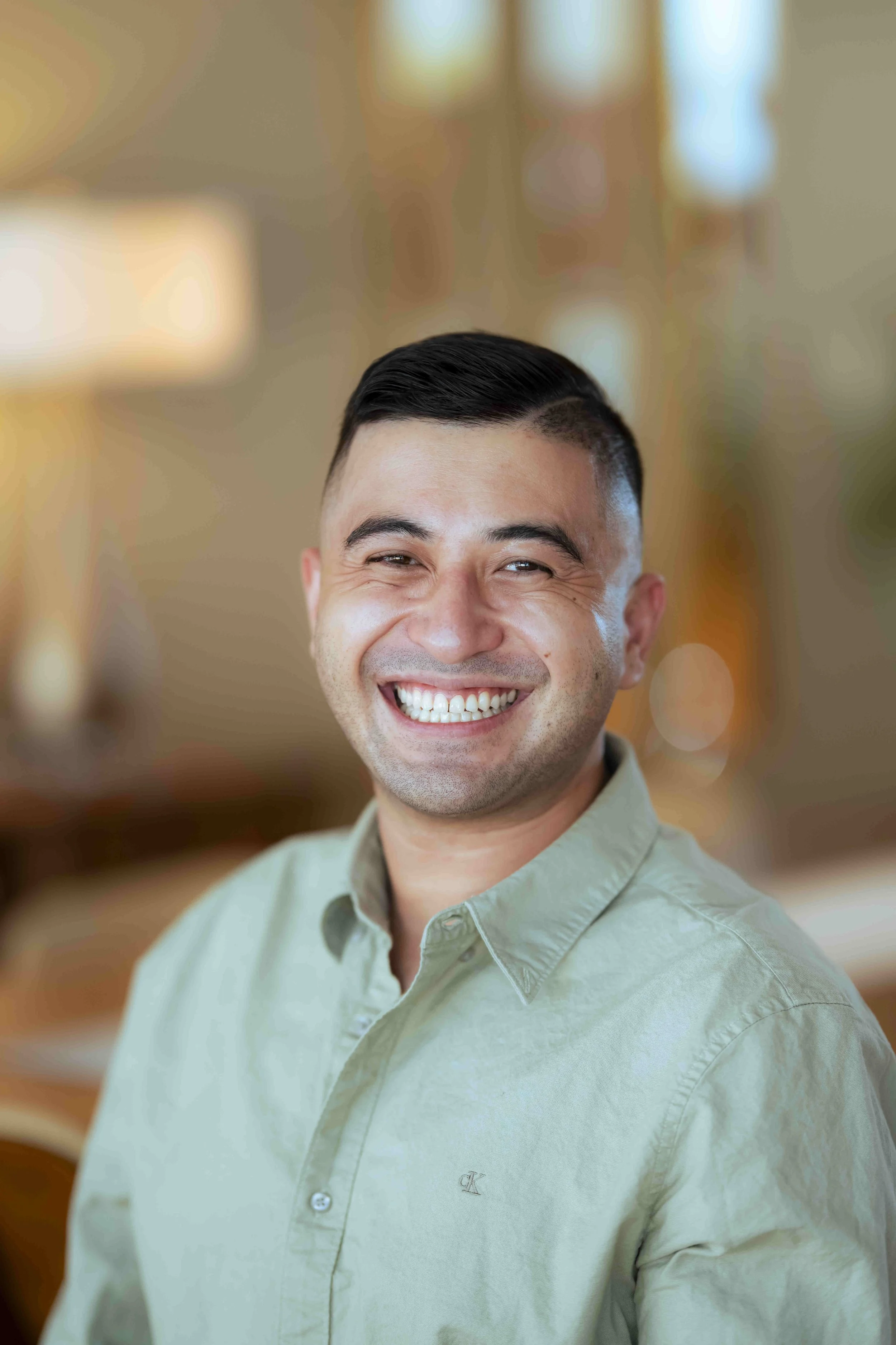 Daniel Mark Hipp smiling, wearing a light-colored shirt, indoors with warm lighting and blurred background.