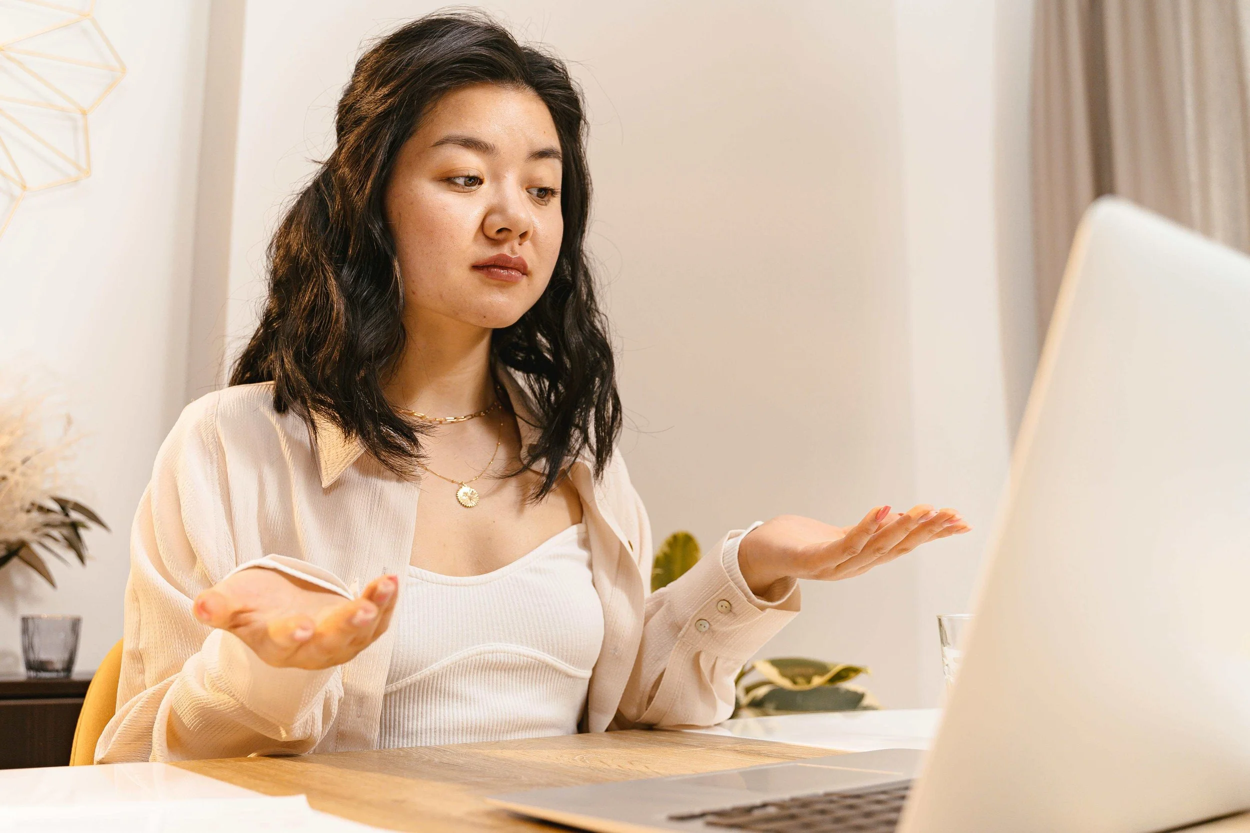 Young woman with dark, wavy hair looking at a laptop, gesturing with her hands in a confused or questioning manner, in a well-lit room with neutral decor.