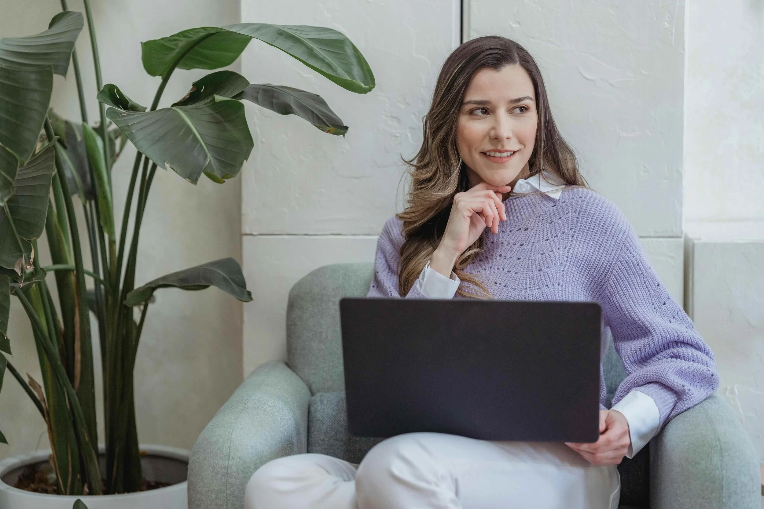 A woman sitting on a light gray armchair with a laptop on her lap, wearing a white shirt and a lavender sweater, smiling thoughtfully while looking to her right, beside a large green houseplant.