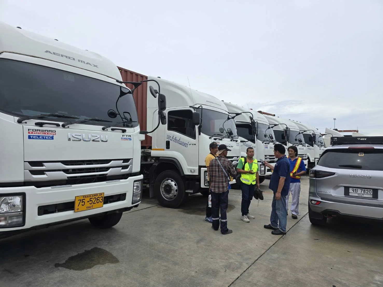A group of people stand and talk near a row of white Isuzu trucks in a parking lot on a cloudy day.