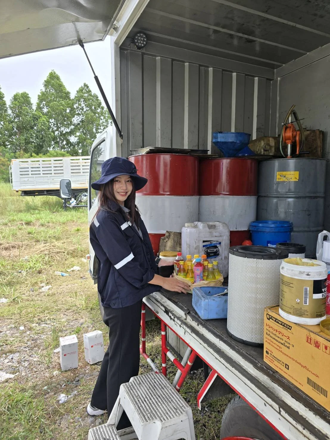 A woman standing inside a mobile food or beverage cart outdoors, smiling at the camera. She is wearing a navy blue hat and uniform. The cart has various supplies including bottles, containers, and equipment, with a grassy field and trees visible in the background.