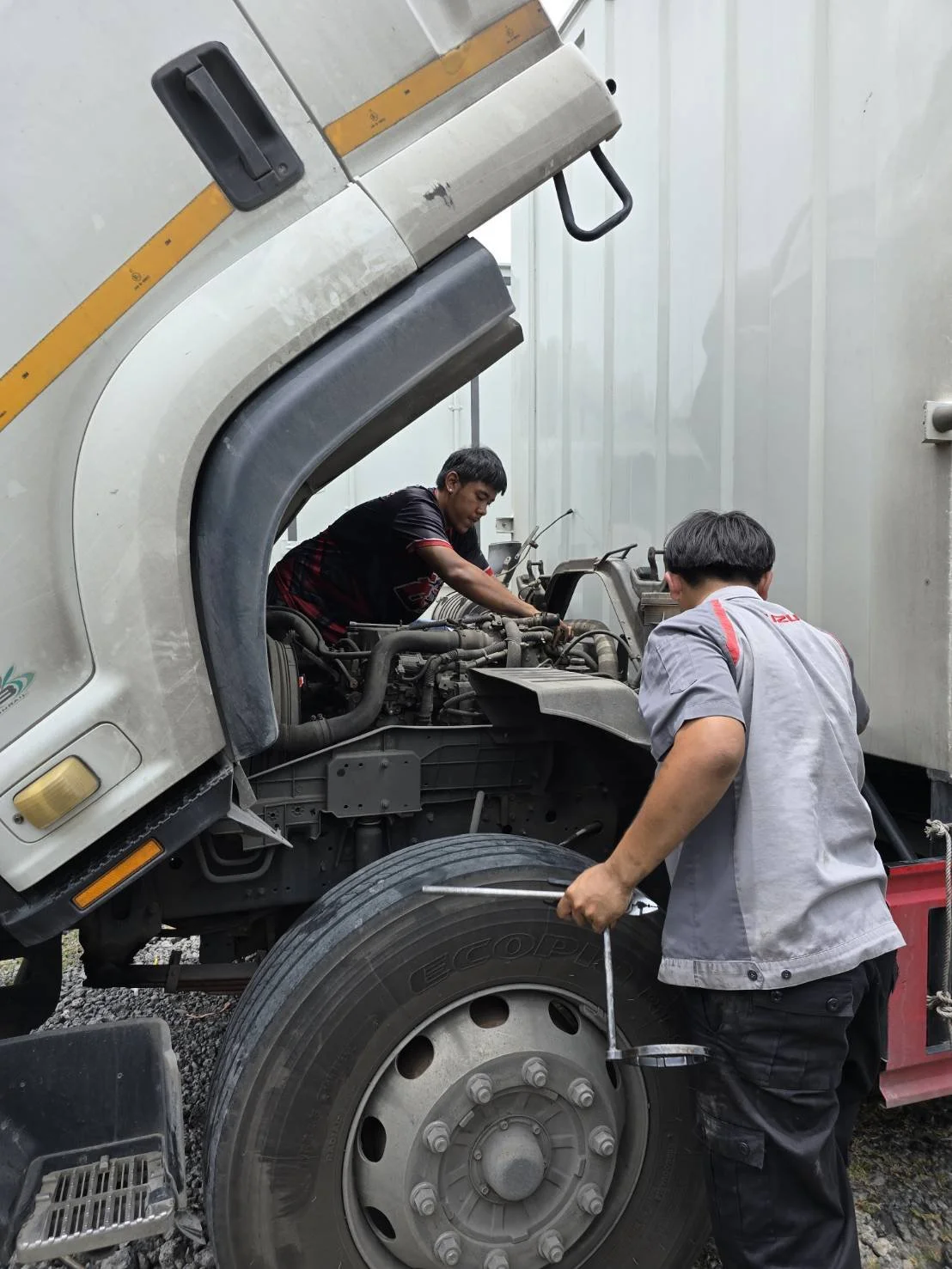 Two men repairing a large truck engine, one is leaning over the engine on the truck, and the other is holding a wrench near the front wheel.