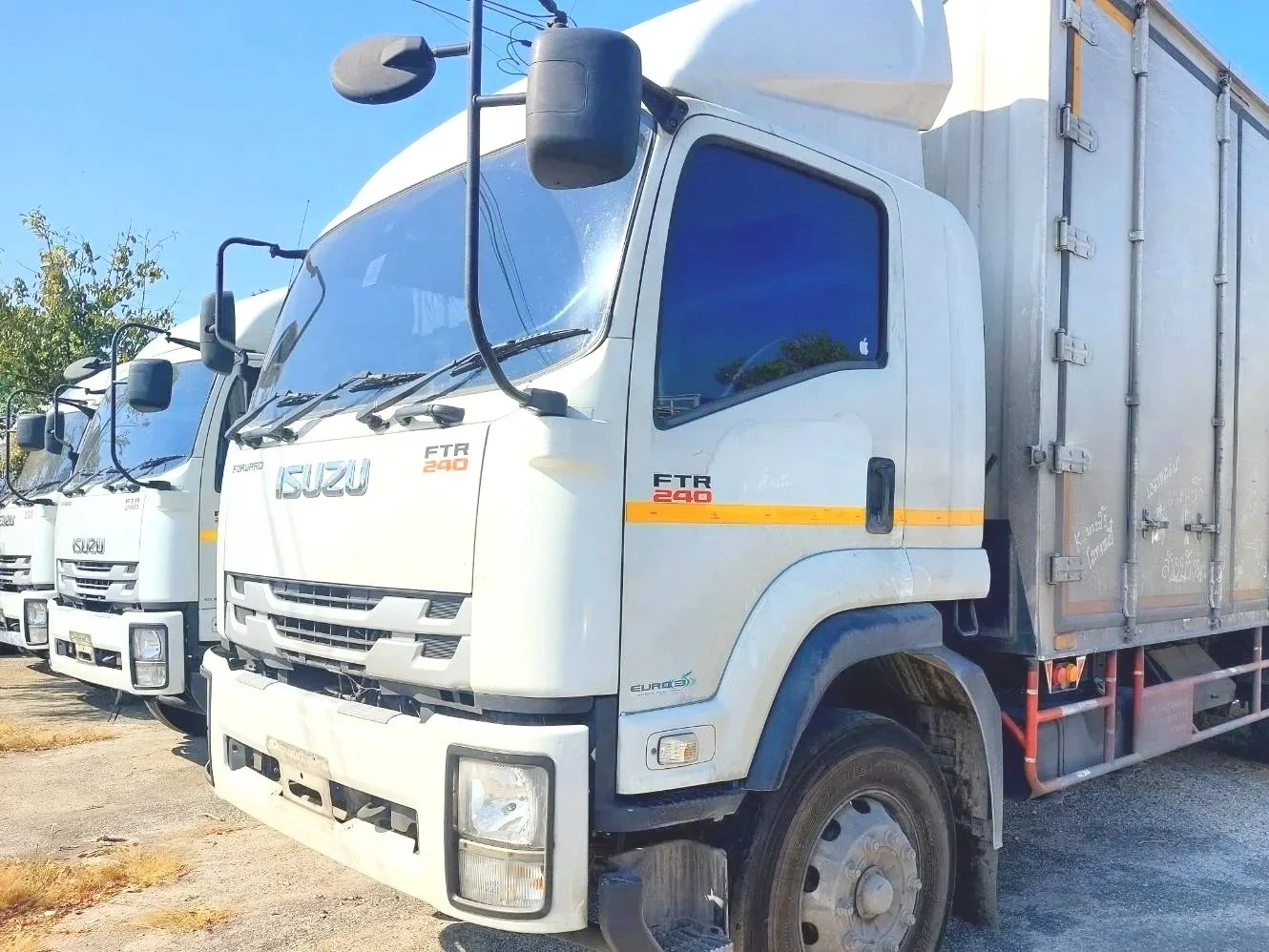 A row of white delivery trucks parked outdoors, with the front truck prominently in view. The trucks have markings including the brand ISUZU and model FTR 240, with a yellow stripe on the side. The background features a clear blue sky and some trees.