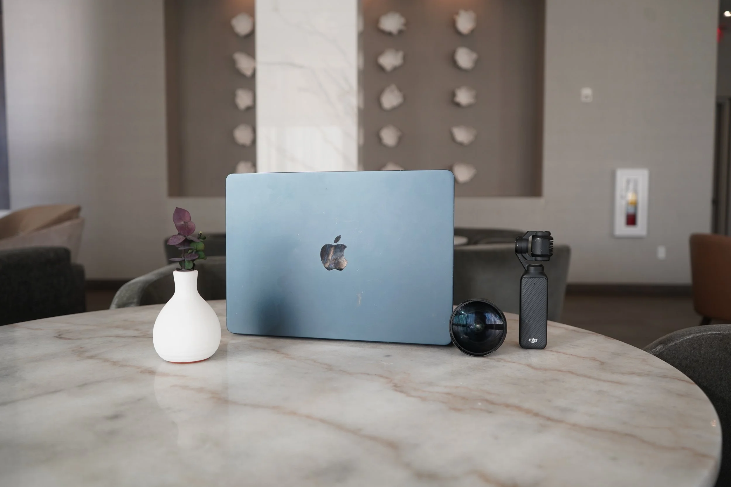 A marble table with a white vase holding purple leaves, a closed blue laptop, a camera lens, and a DJI handheld gimbal, with a blurred background of a modern interior wall with decorative pieces and a fire extinguisher.
