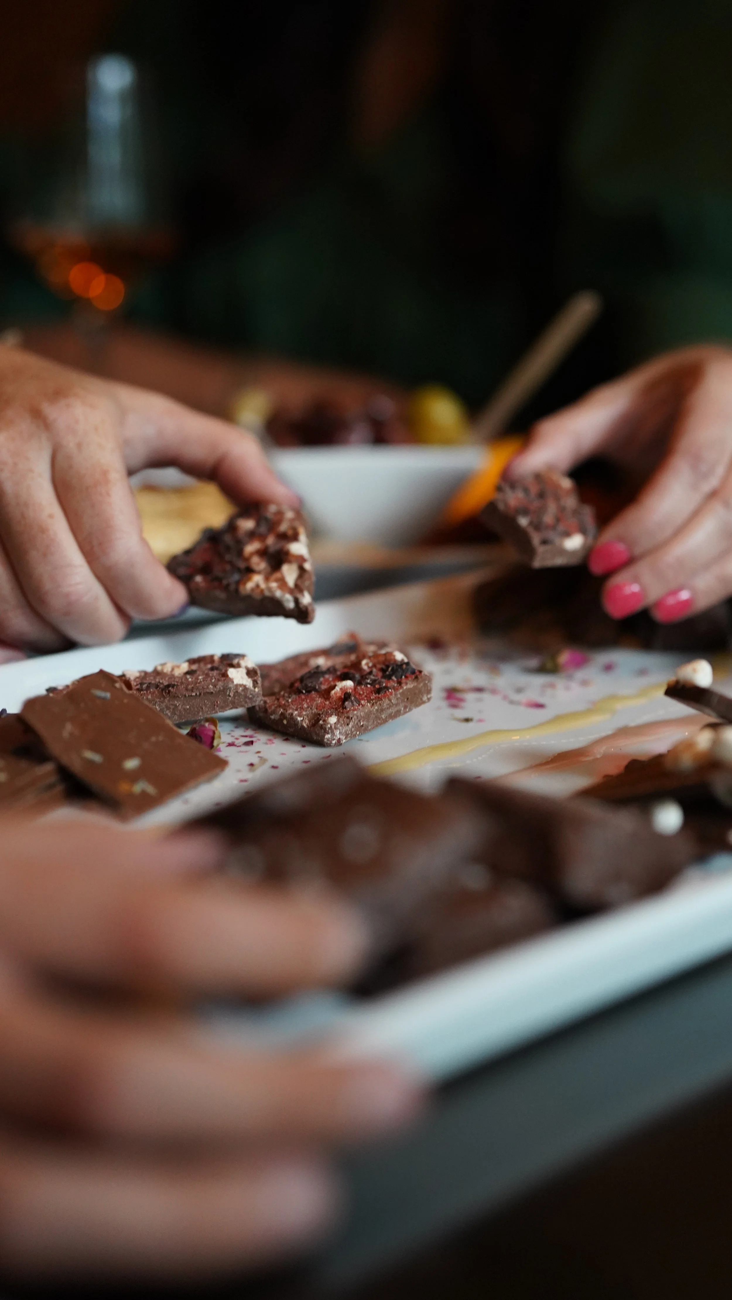 Hands breaking pieces of assorted chocolate on a white plate in a close-up shot.