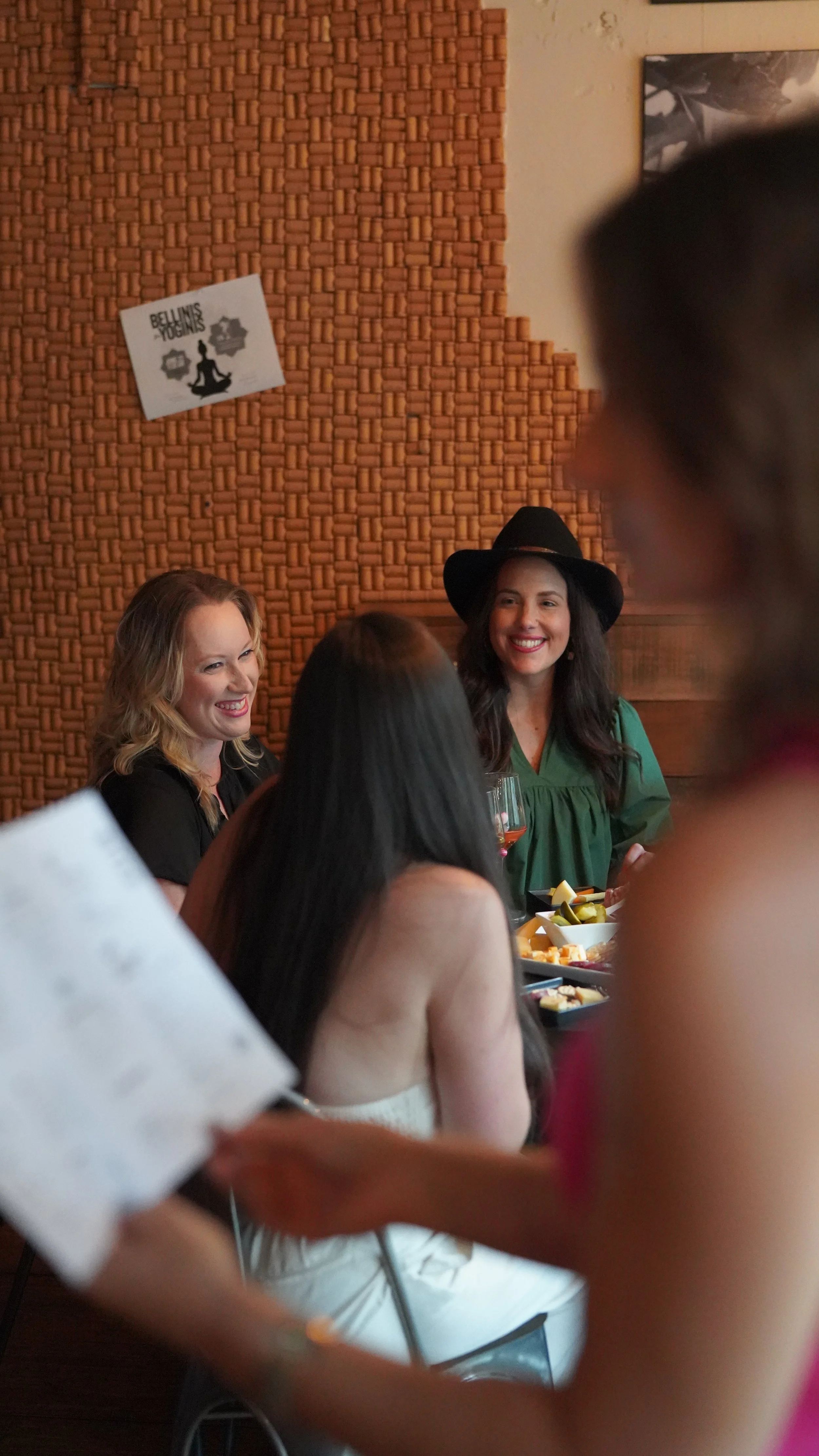 Women enjoying a social gathering, with one woman wearing a black hat and green top smiling, and others engaged in conversation, with a brick wall and a small sign in the background.