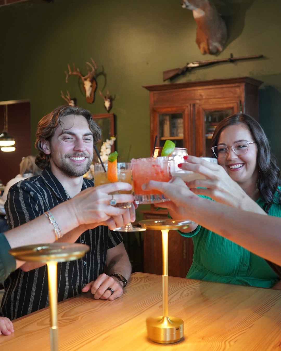 People enjoying cocktails around a table in a cozy bar or restaurant with deer antlers and hunting rifles mounted on the green wall behind them.