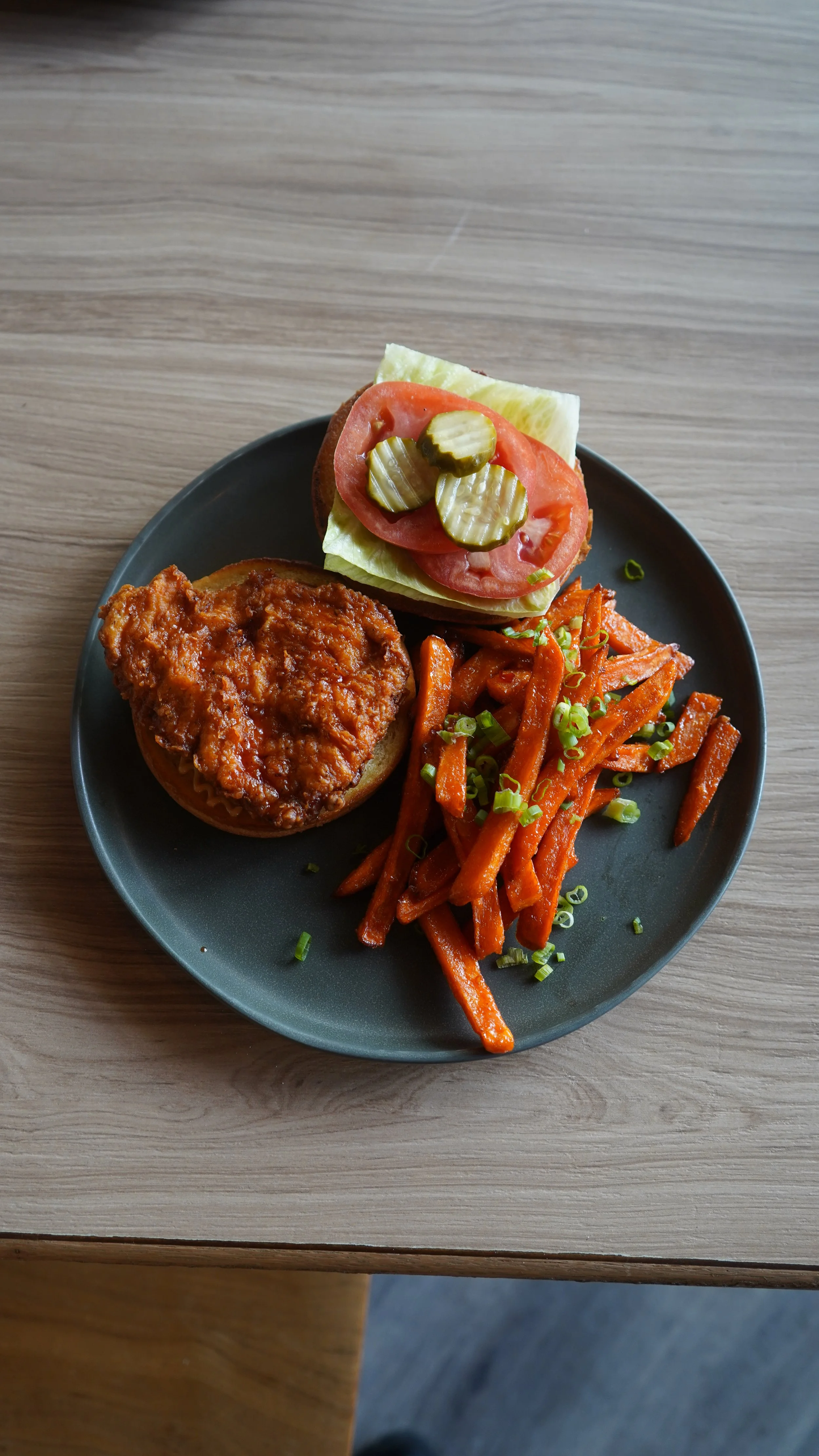 Plate with a serving of chili over a bun, a small sandwich with lettuce, tomato, and pickles, and a side of glazed carrots garnished with chopped green onions, on a wooden table.