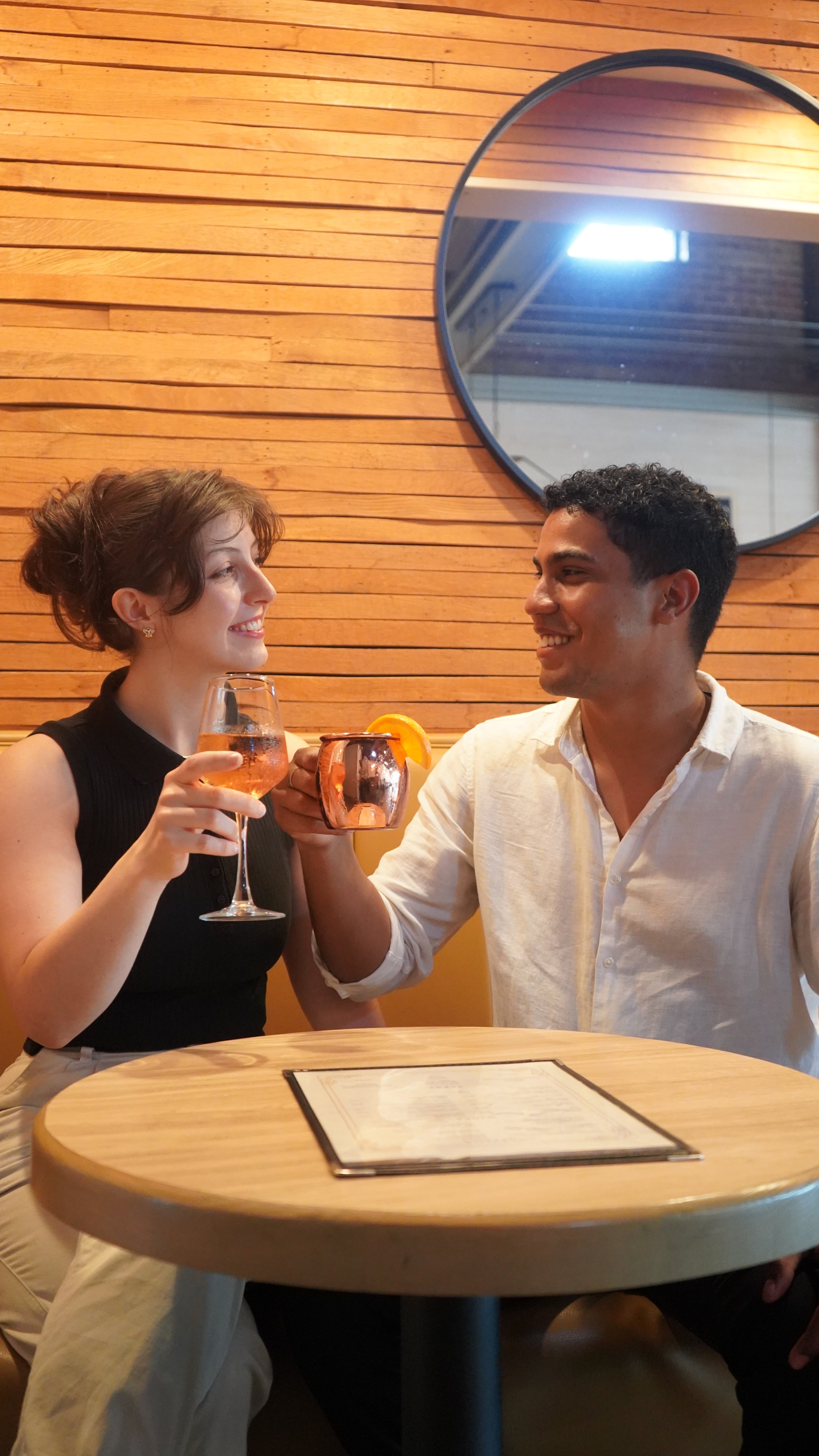 A woman and man sitting at a round wooden table in a restaurant, sharing a toast with drinks, smiling at each other.