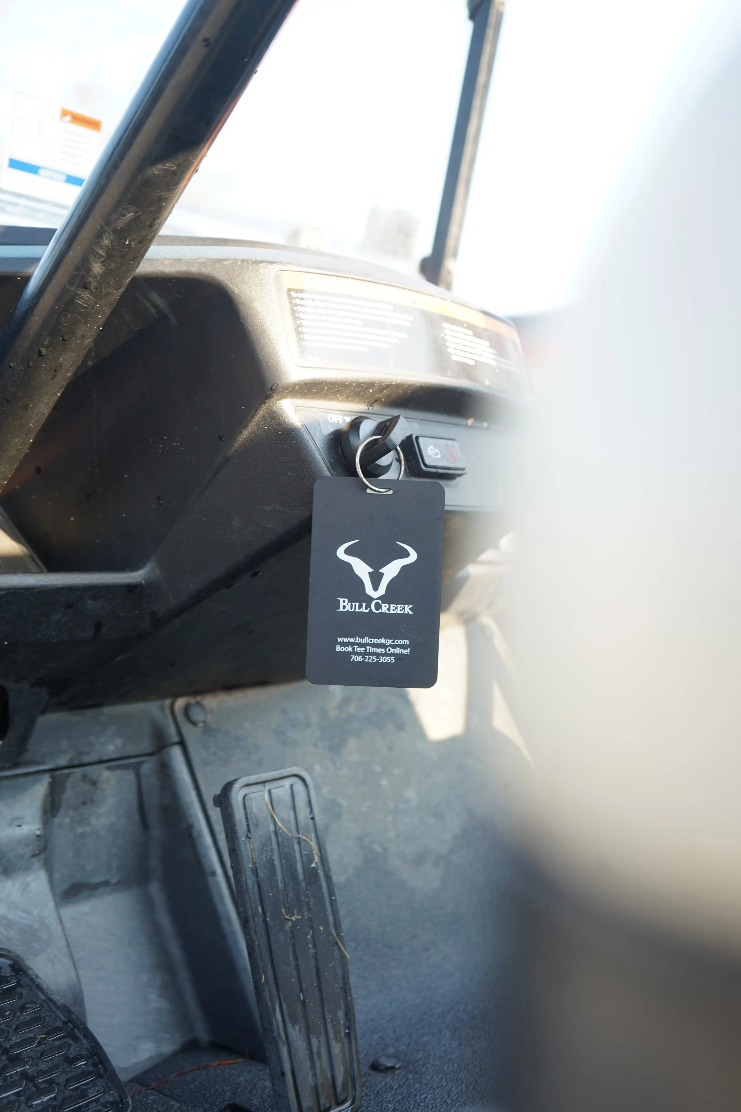 The interior of a vehicle showing a key in the ignition with a black Bull Creek tag hanging from it, and part of the dashboard and driver's side foot pedal area.