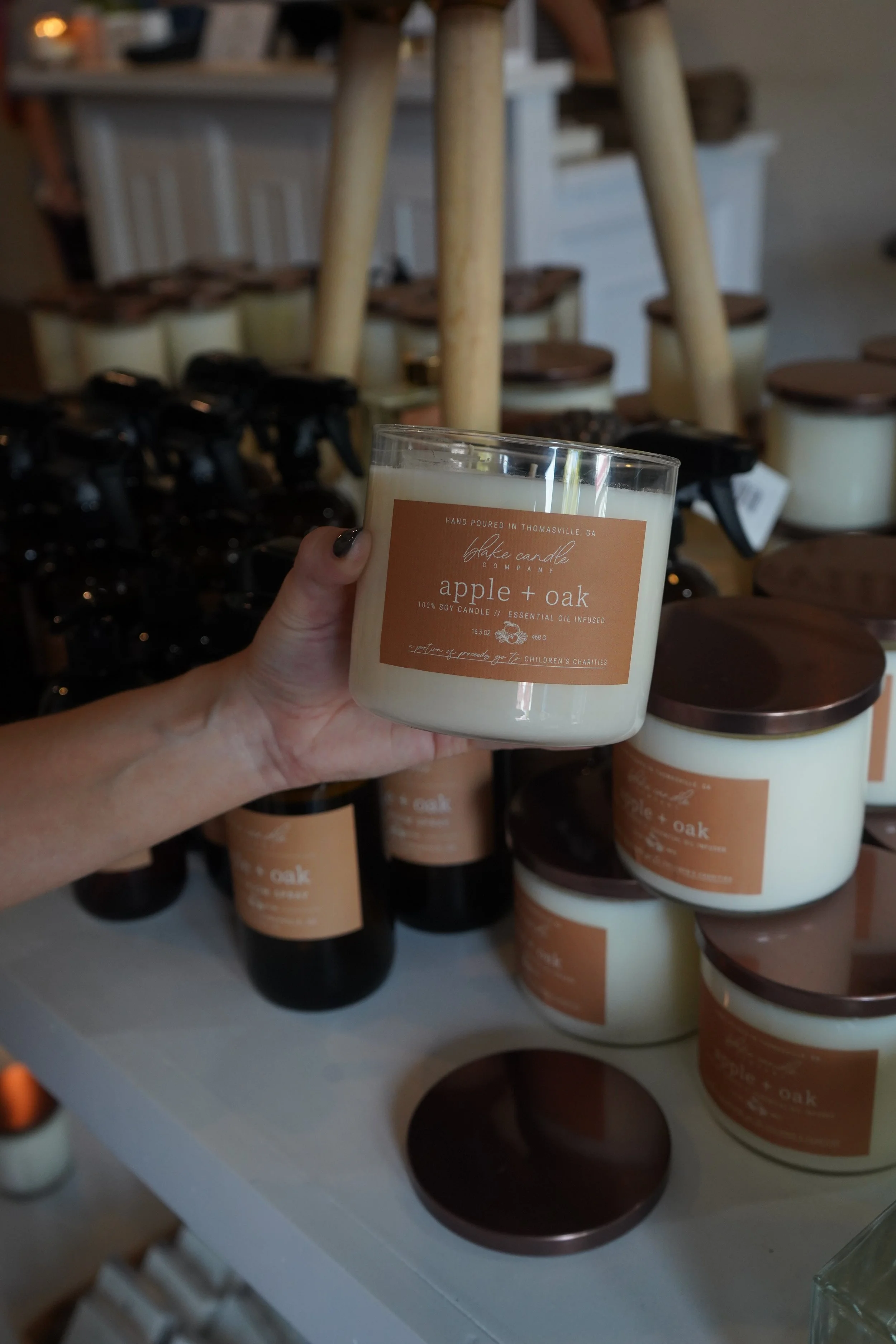 A hand holding a white candle with a label that reads 'apple + oak'; in the background, there are stacked candles and products on a display table.