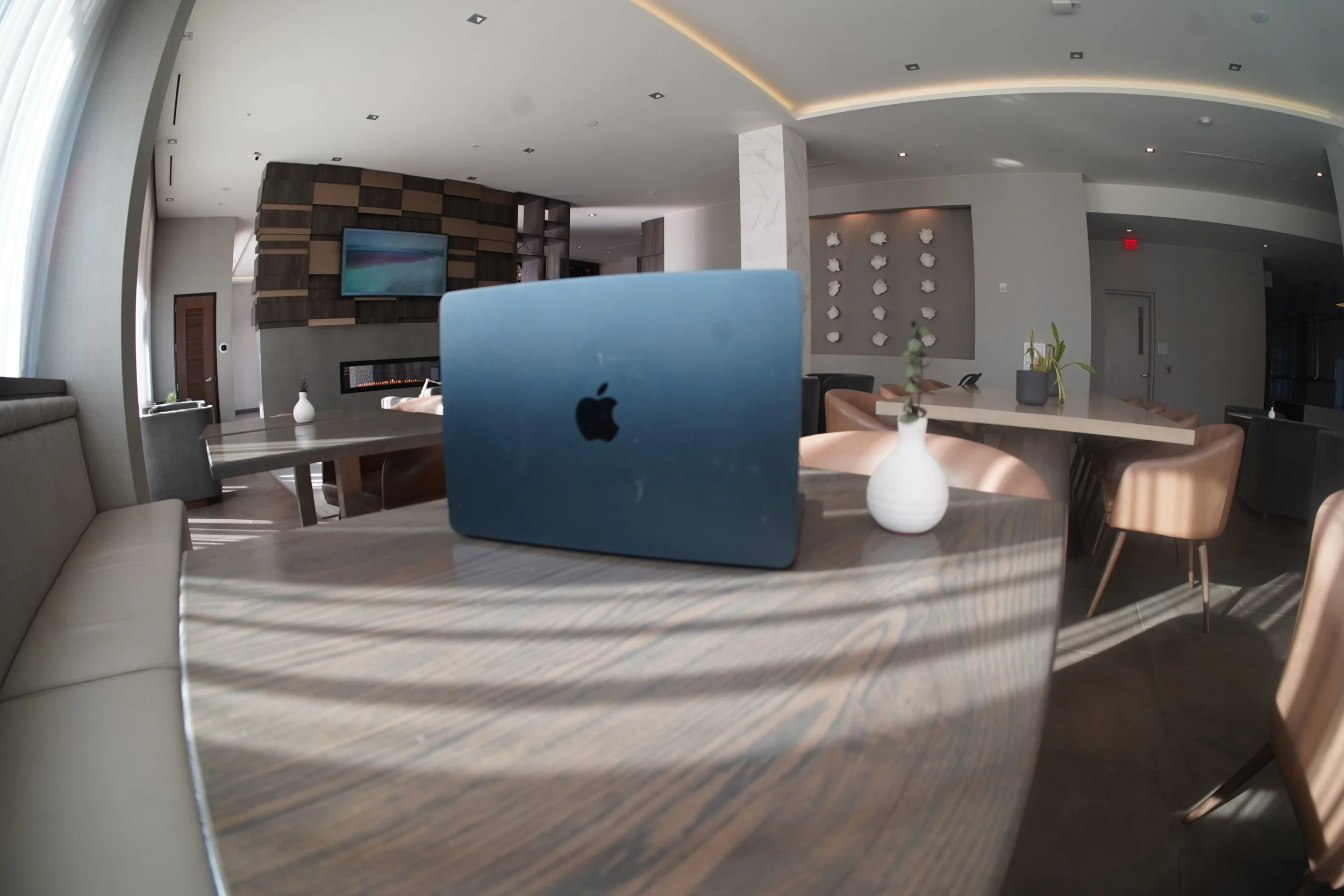 Modern, well-lit restaurant or lounge with wooden tables, beige and black chairs, decorative vases, and a wall-mounted TV.