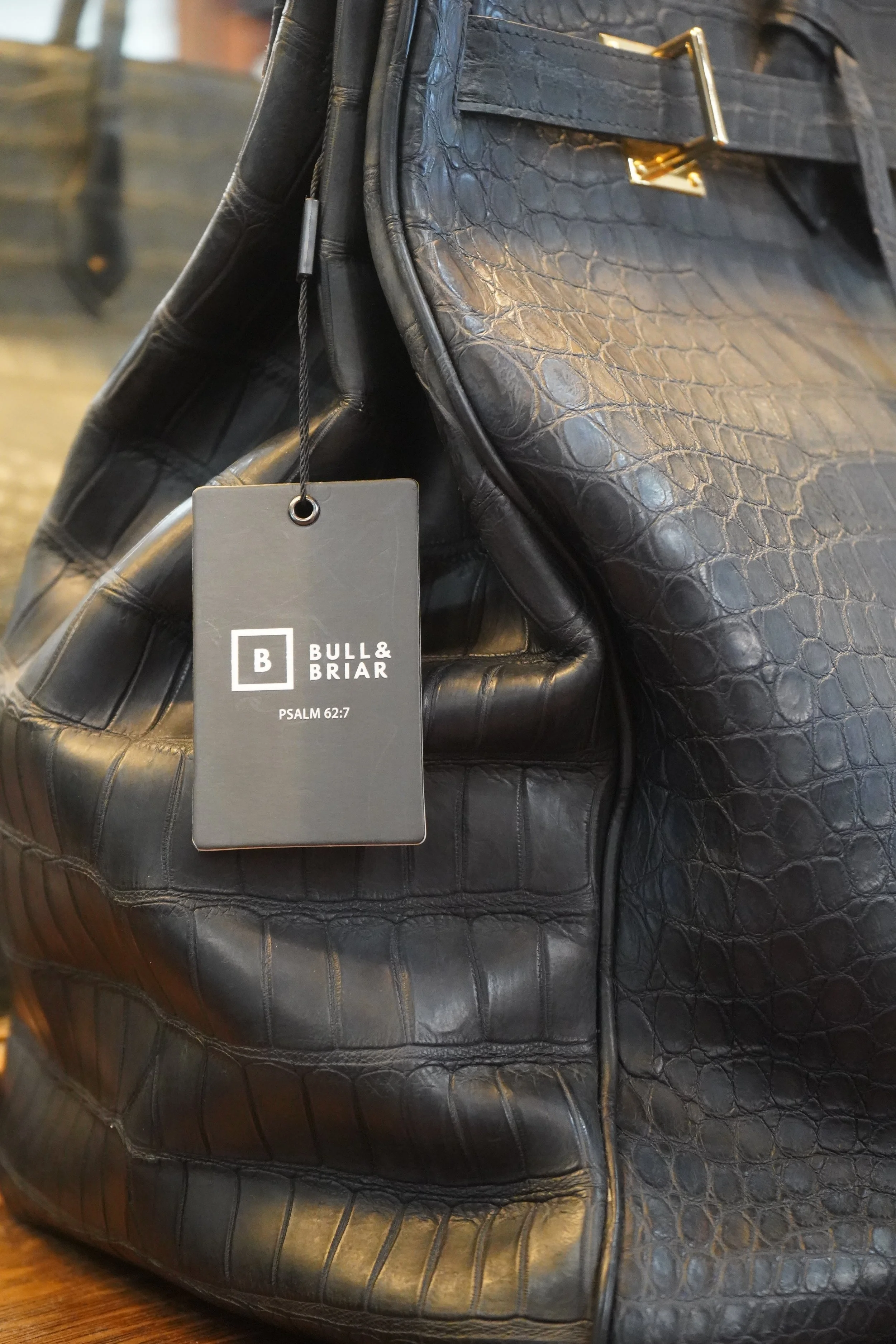 Close-up of a black luxury bag with a BULL & BRIAR tag attached, featuring textured crocodile-pattern leather and gold hardware on a wooden surface.