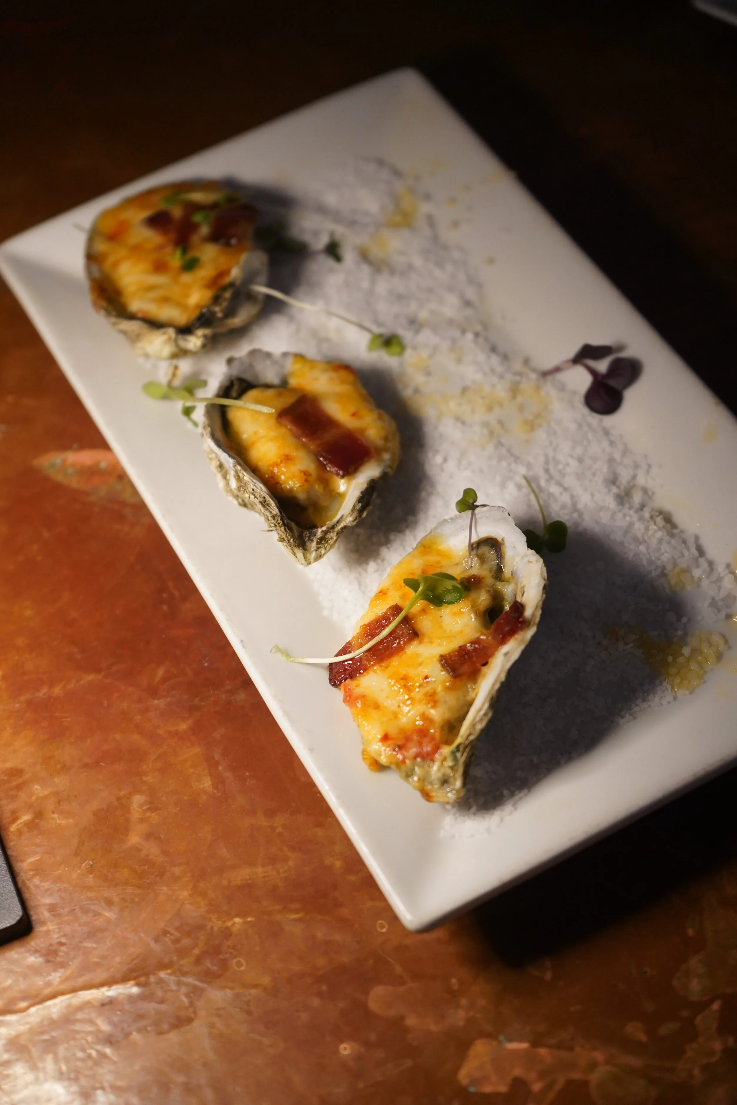 Oysters Rockefeller garnished with microgreens on a white plate with powdered seasoning and small spoonfuls of sauce.