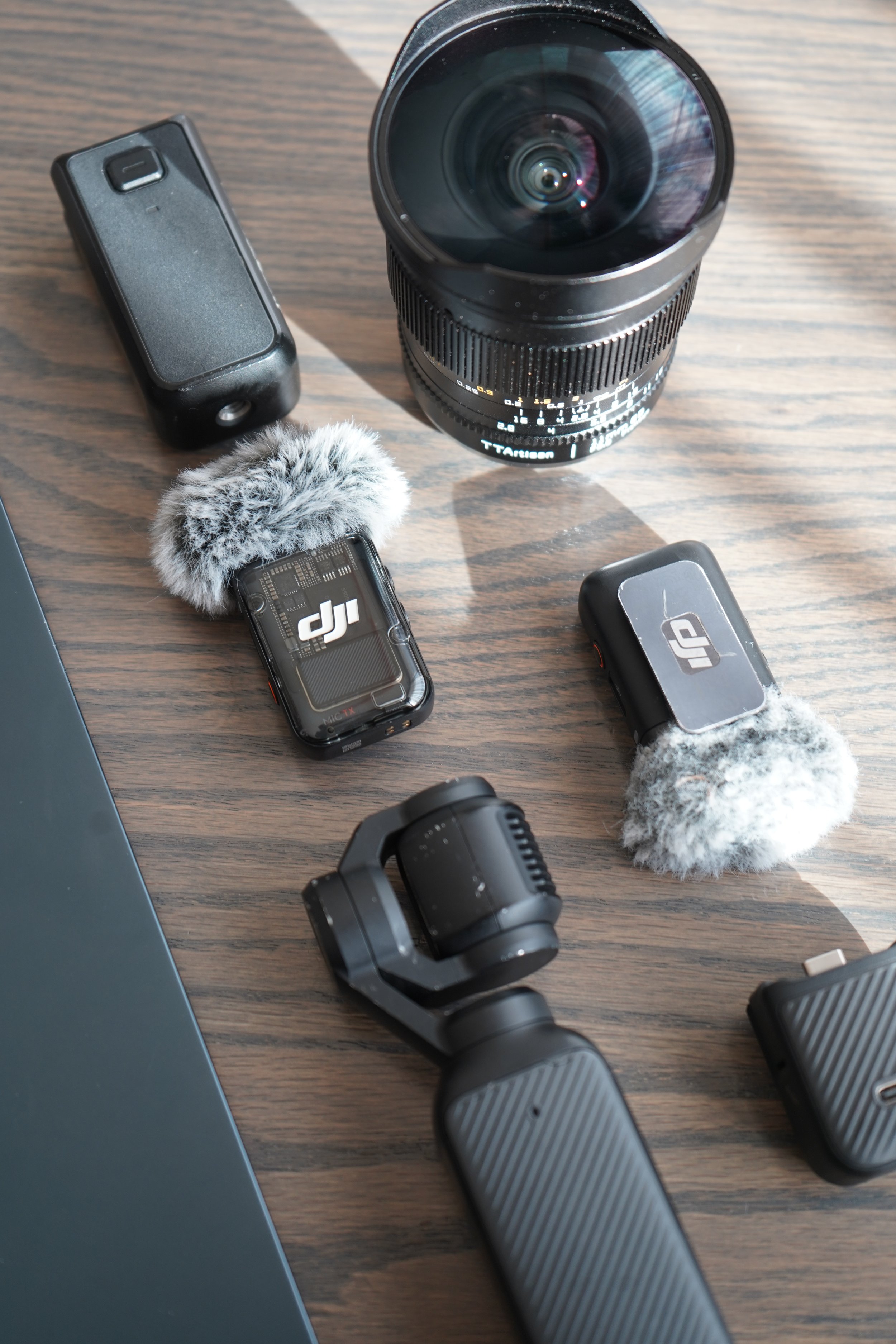 Photography equipment on a wooden surface, including a camera lens, a gimbal stabilizer, remote controls with furry windshields, and other accessories.