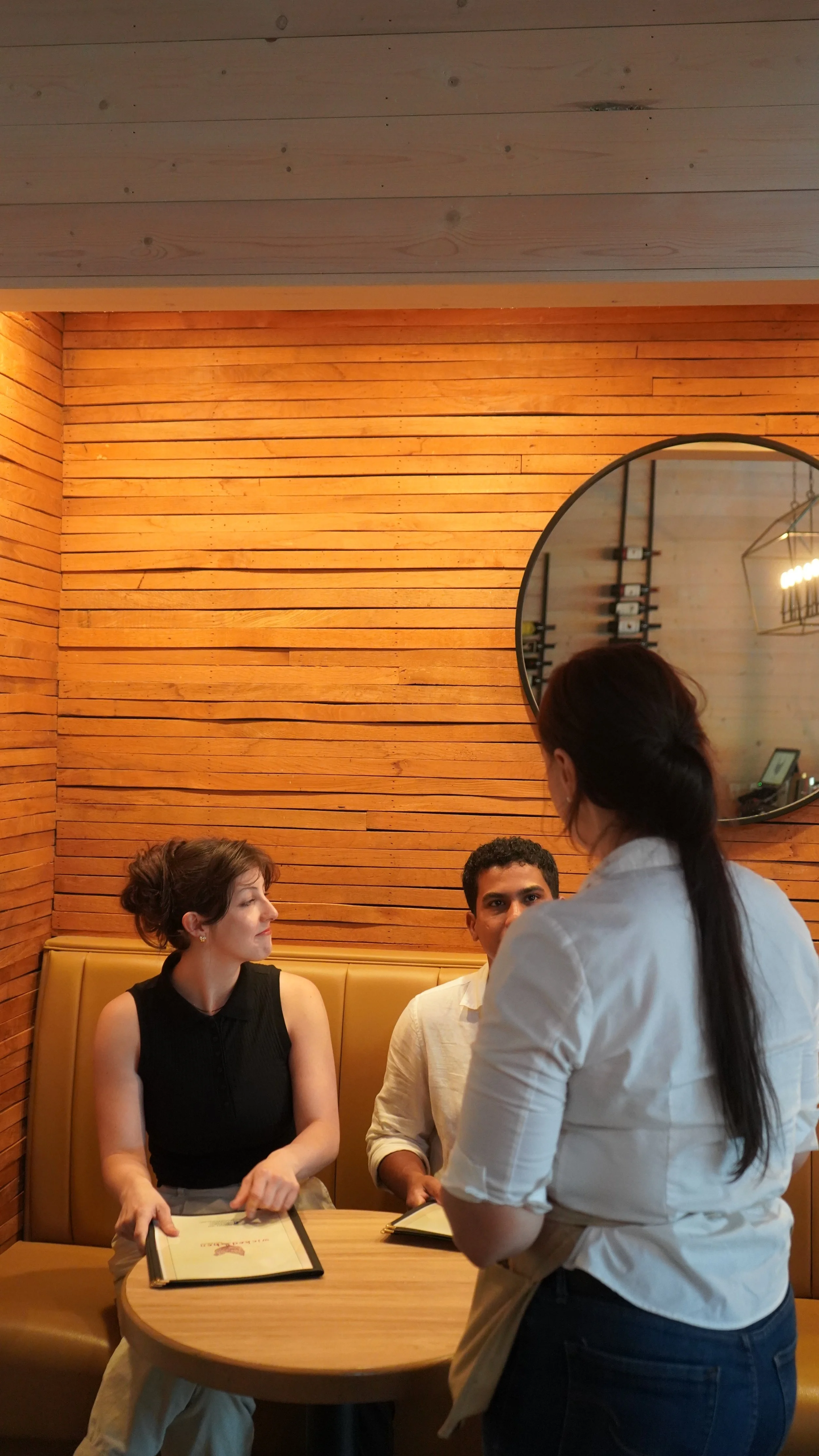 A woman standing and talking to a man and woman sitting at a round table in a restaurant with wood-paneled walls and a mirror.