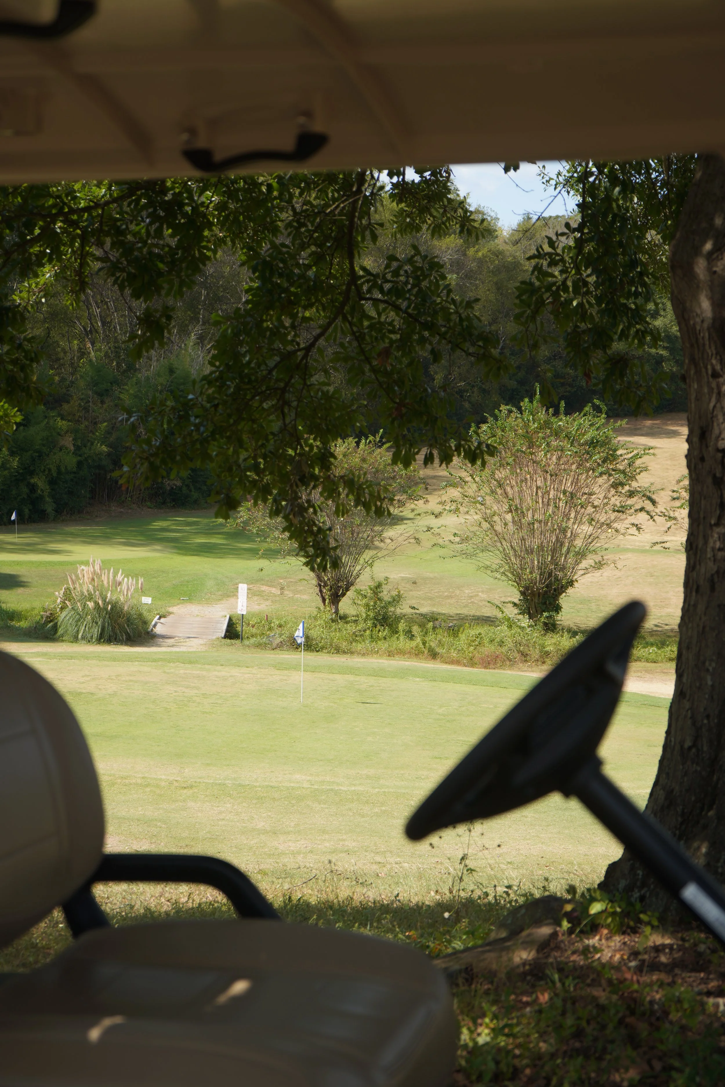 View of a golf course through the windshield of a golf cart, with trees and a flag on a hole