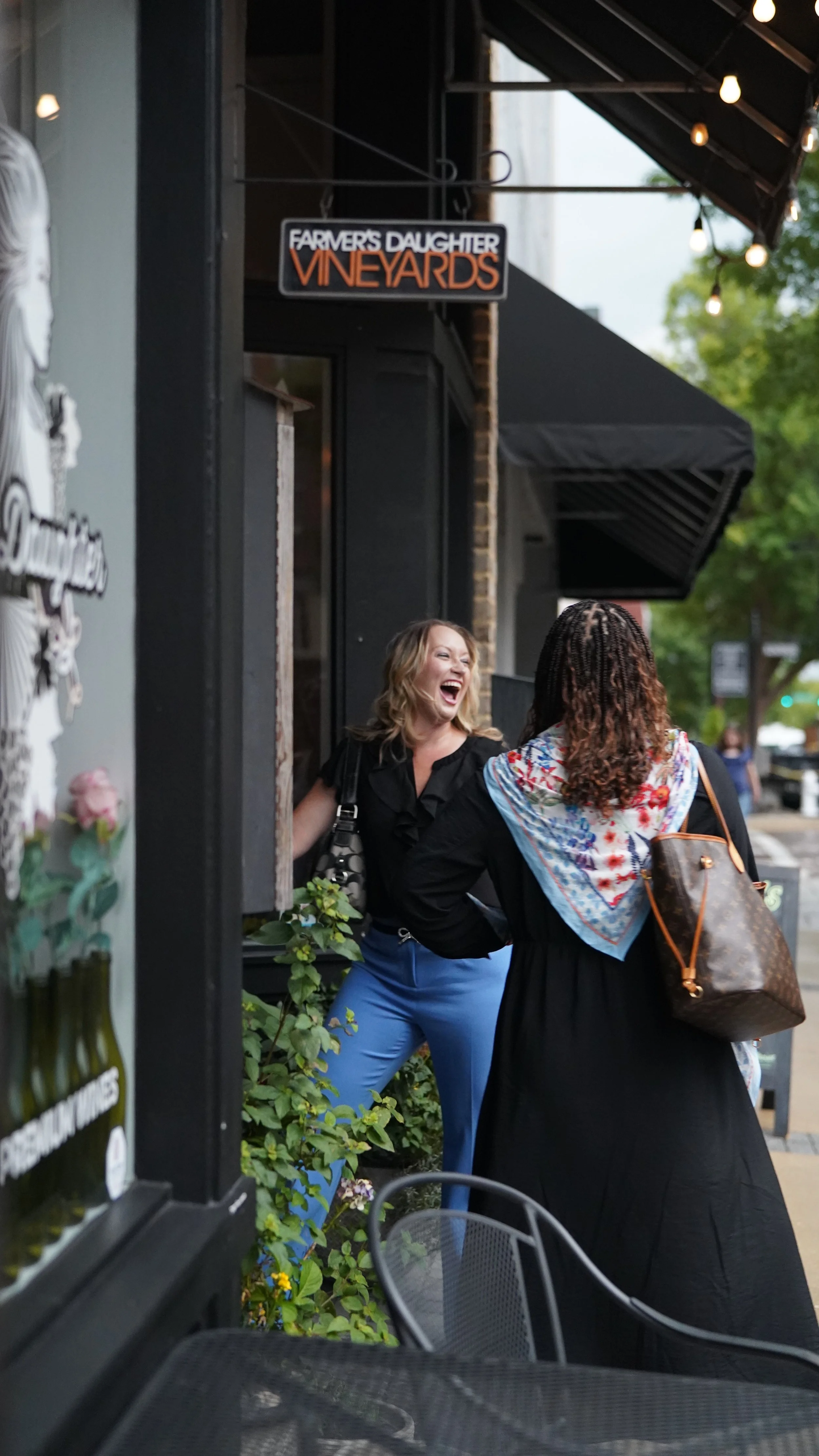 Two women smiling and laughing on a sidewalk outside a business named 'Farmer's Daughter Vineyards.' One woman has blonde hair and is wearing a black top and blue pants, while the other has curly hair, a colorful scarf, and a large handbag.