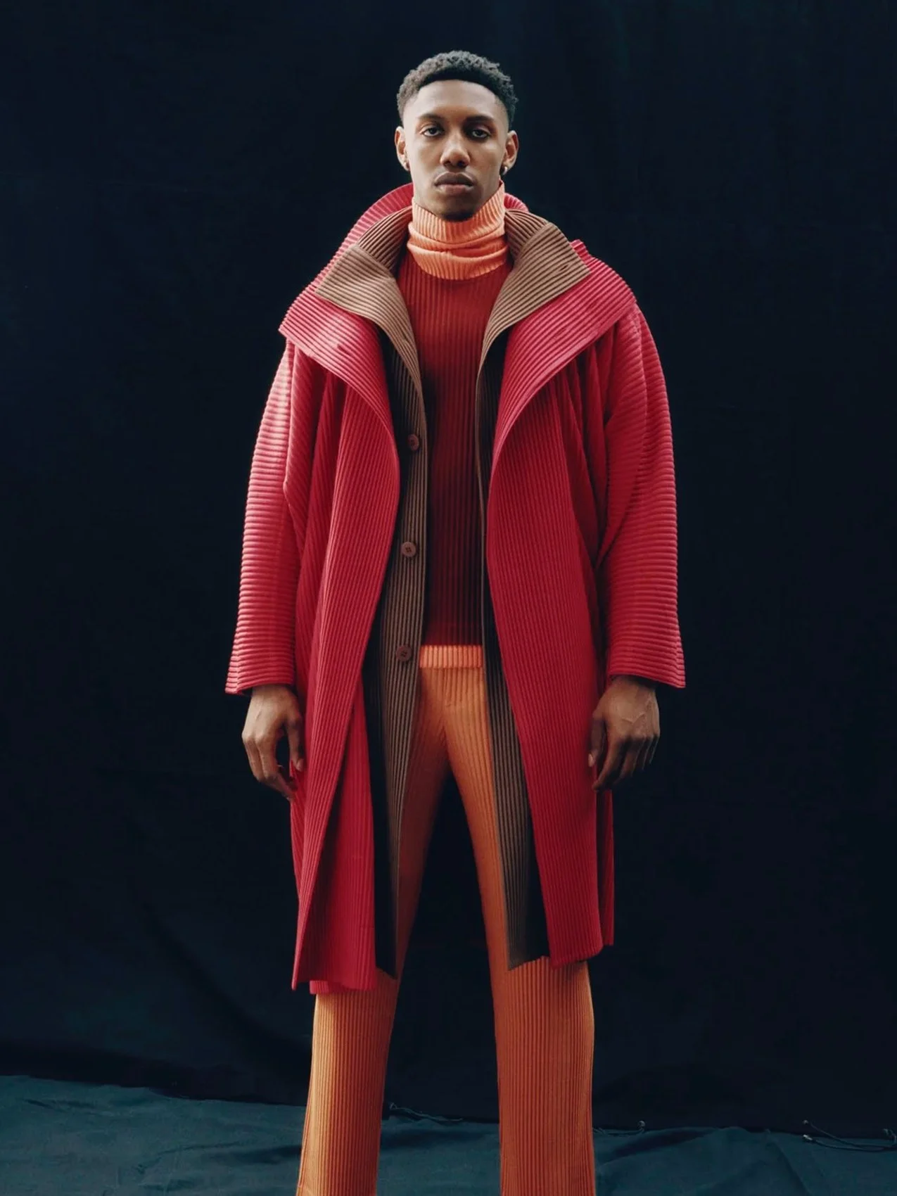 A young man standing against a black background, wearing a multicolored, textured coat and matching striped pants, with a turtleneck sweater.