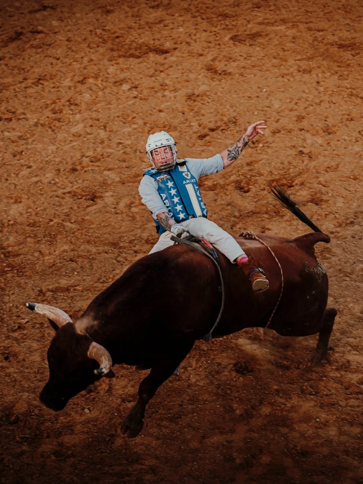 A person wearing a helmet and protective gear riding a charging bull with dust flying up on a dirt arena.
