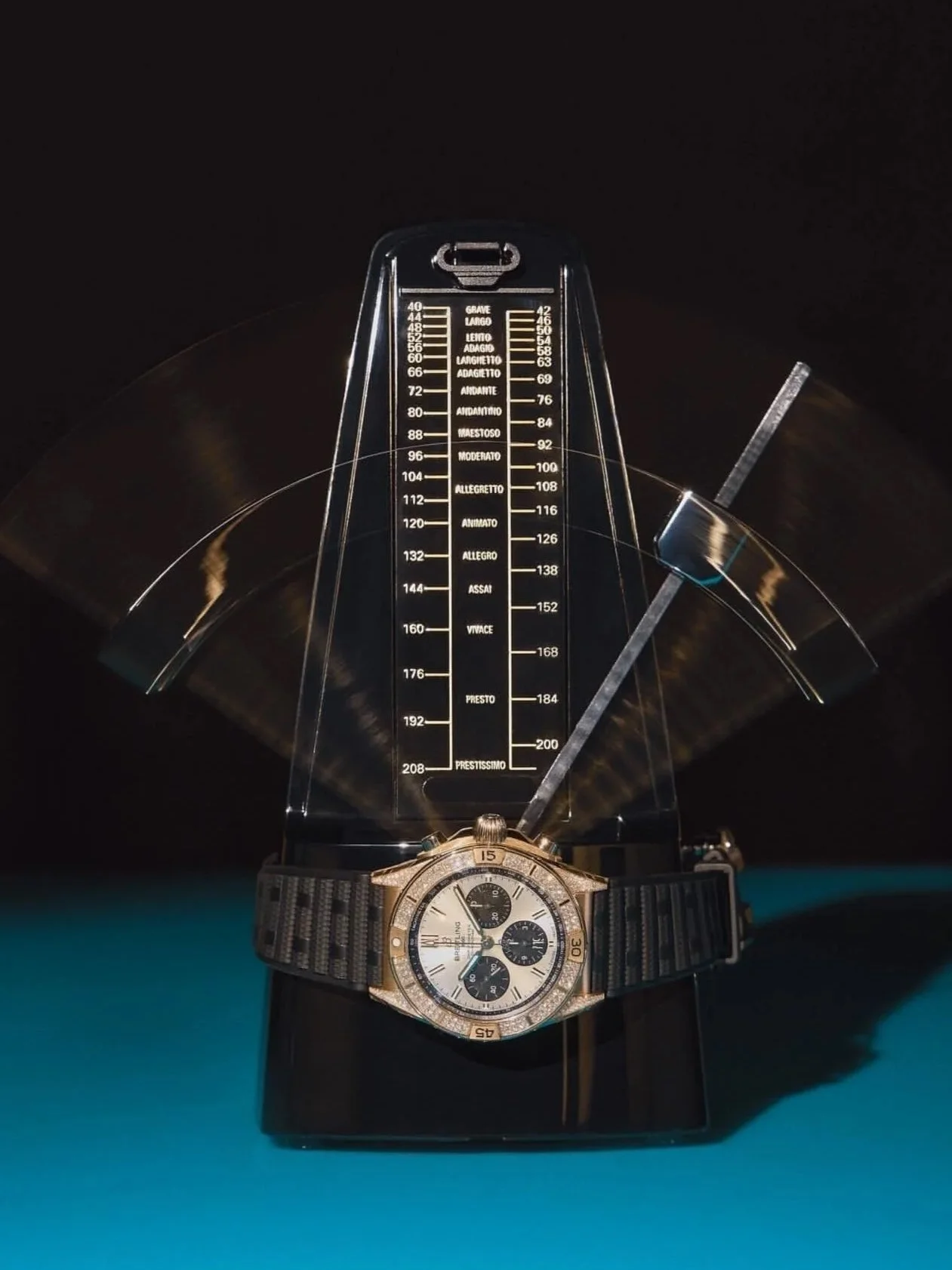 A wristwatch resting on a black surface with a large black and silver, chronograph style dial and metal bracelet, placed in front of a caliper measuring thick transparent plastic measurer with scale.