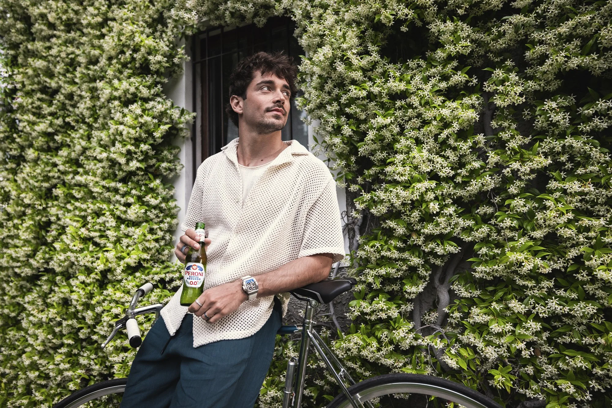 Man with dark curly hair wearing a cream-colored mesh shirt and teal pants, holding a Peroni beer bottle, leaning on a bicycle, standing against a wall covered with white flowering vines, looking to his left.