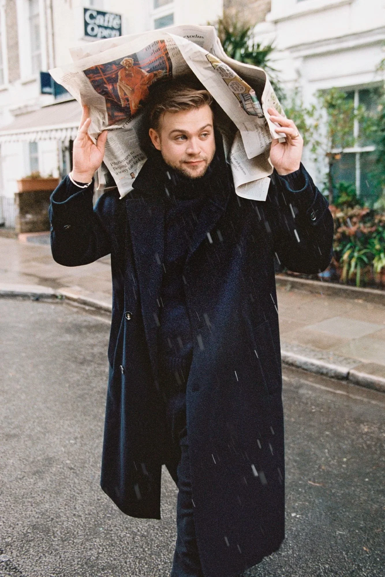 A man with short hair and facial hair carrying a newspaper on his head while walking on a rainy street.