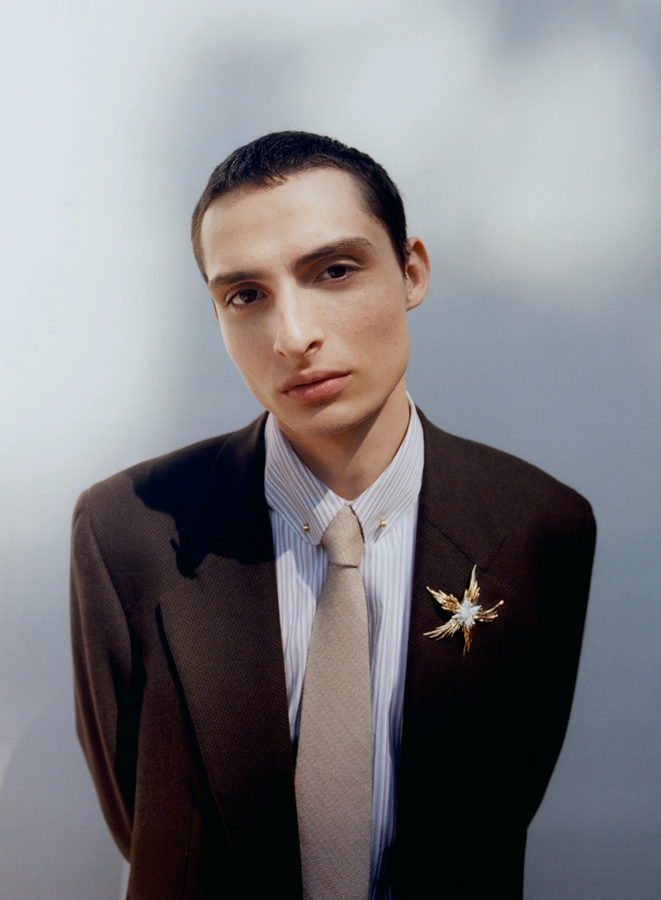 A young man with fair skin and short dark hair wearing a brown suit, white striped shirt, beige tie, and a gold and white brooch on his lapel, standing against a plain light gray background.