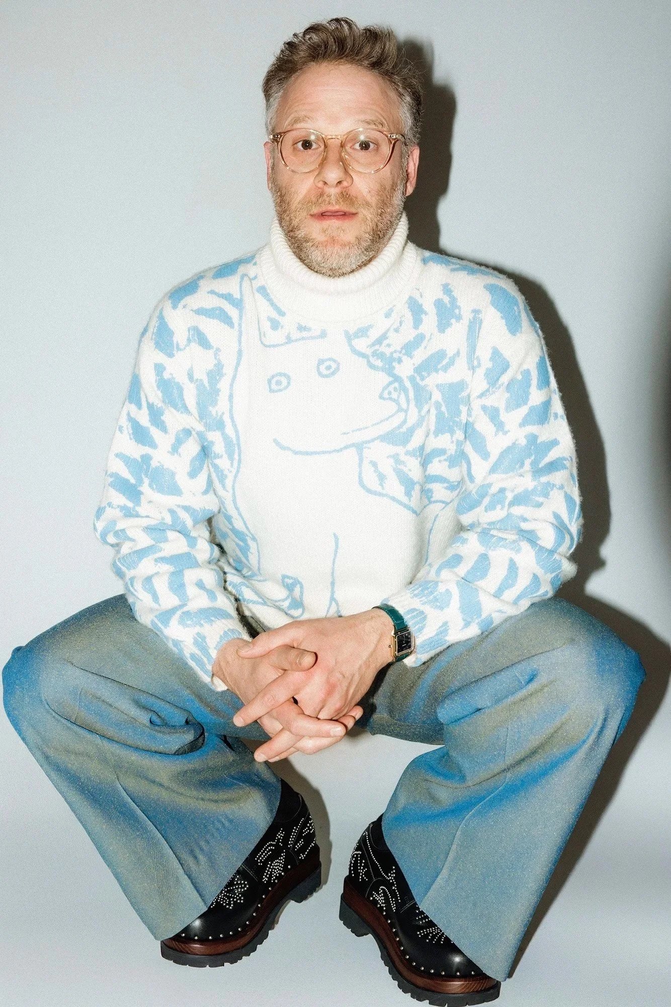 Seth Rogen squatting for a photoshoot wearing a light blue sweater