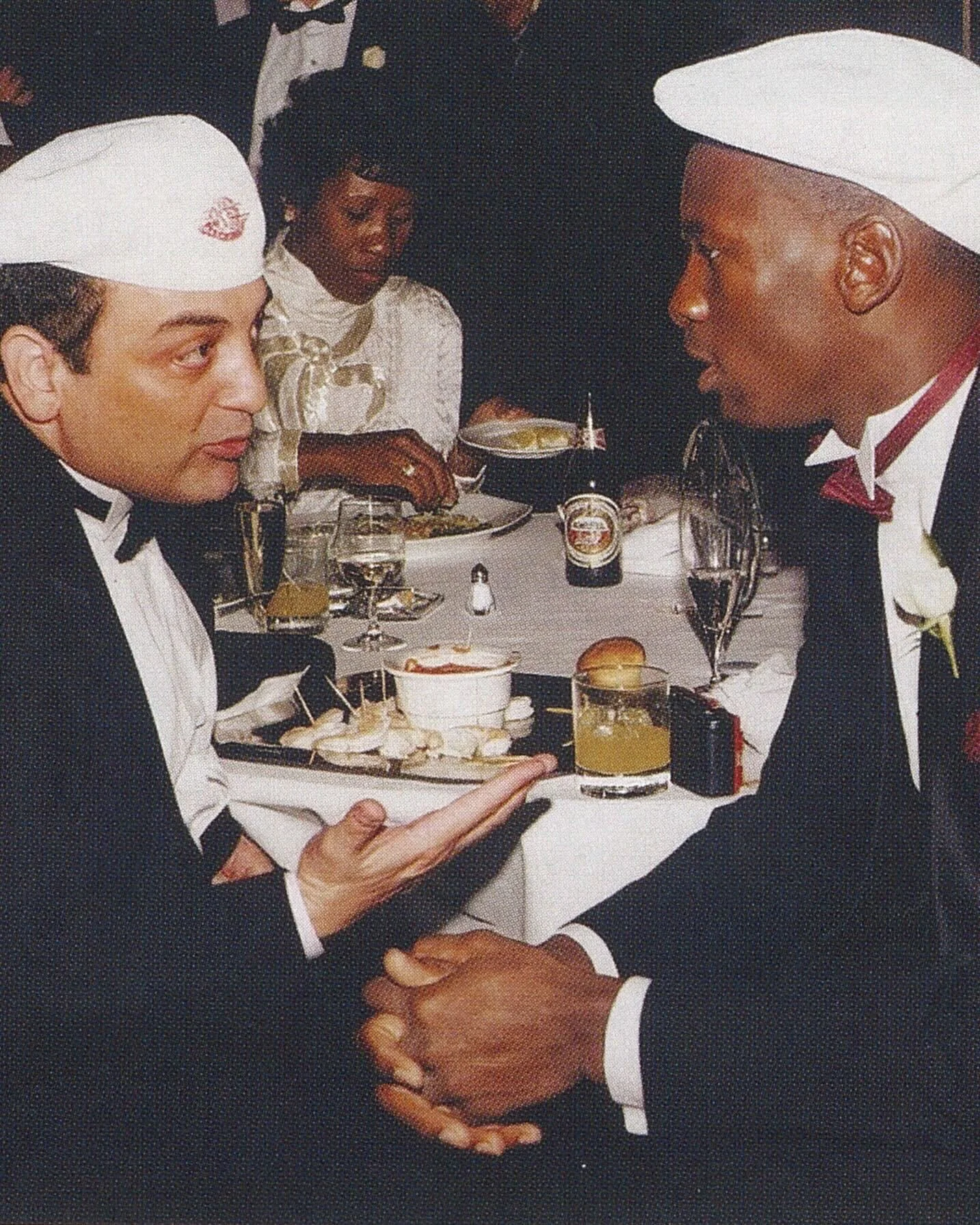 Two men dressed in tuxedos and white hats holding hands and looking at each other at a formal dinner event, with a woman in a white dress seated in the background.