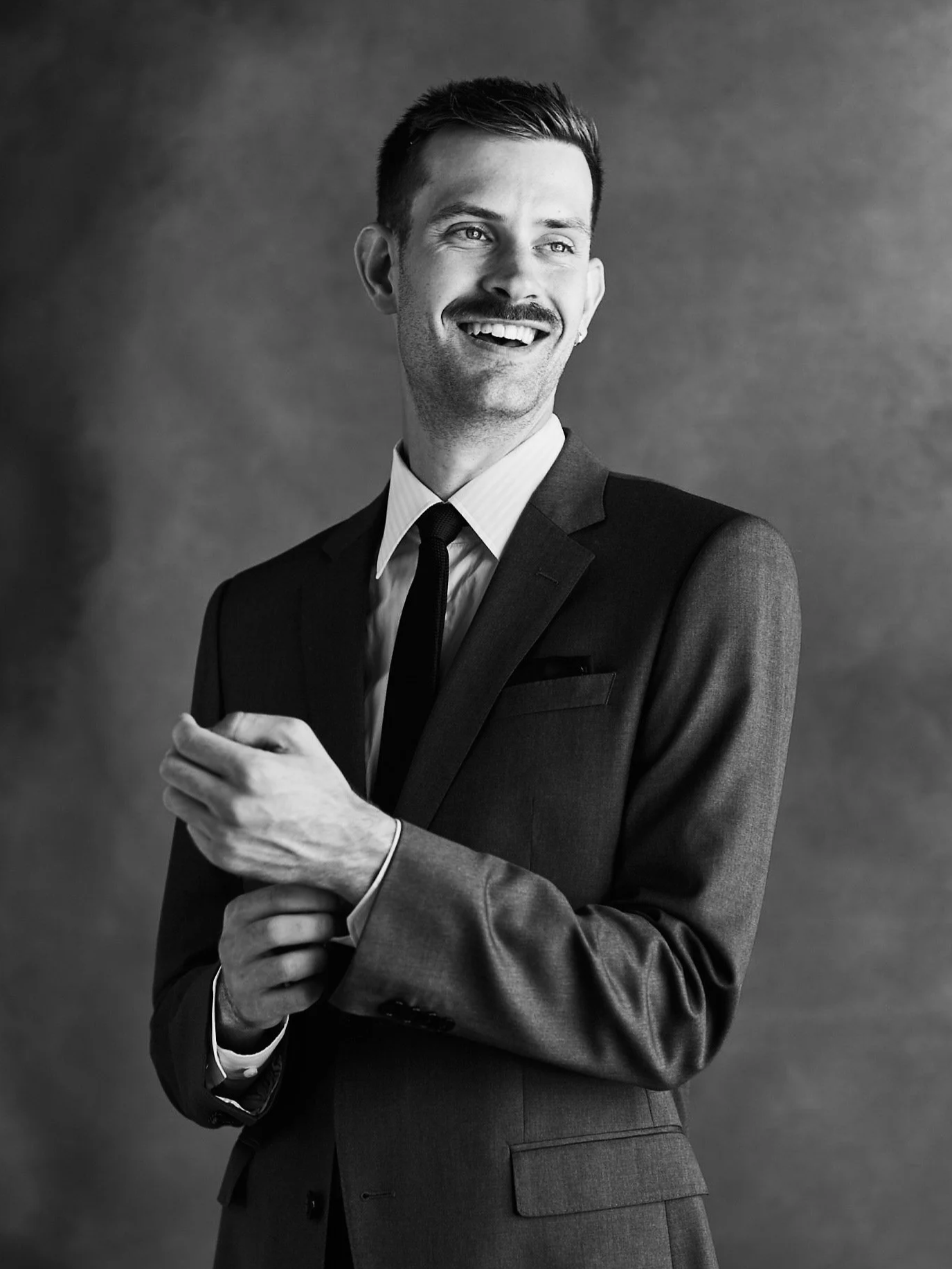 A black and white photo of a man in a suit smiling and looking to his right, holding his sleeve with his left hand.