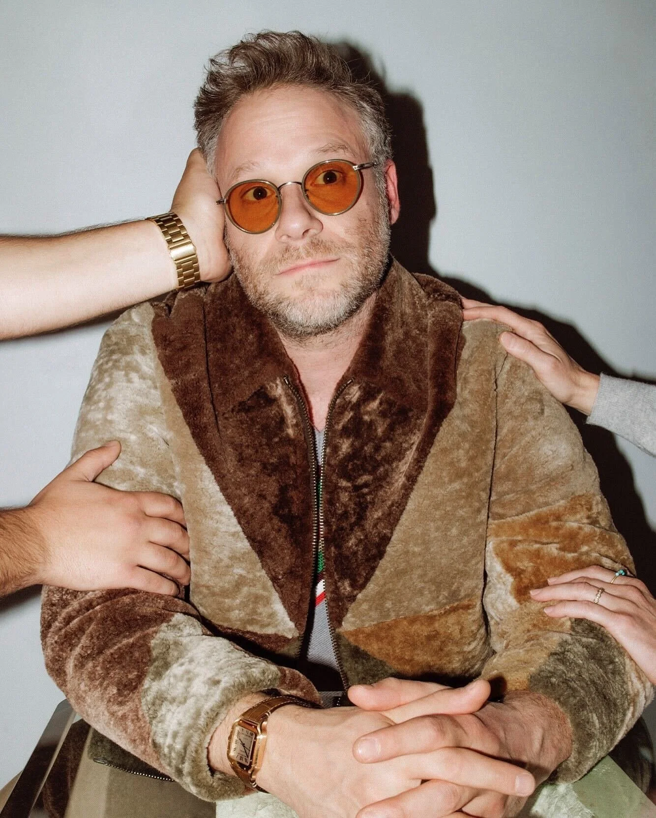 A middle-aged man with gray hair and beard wearing orange sunglasses and a brown velvet patchwork jacket, sitting with arms crossed and hands resting on a table.
