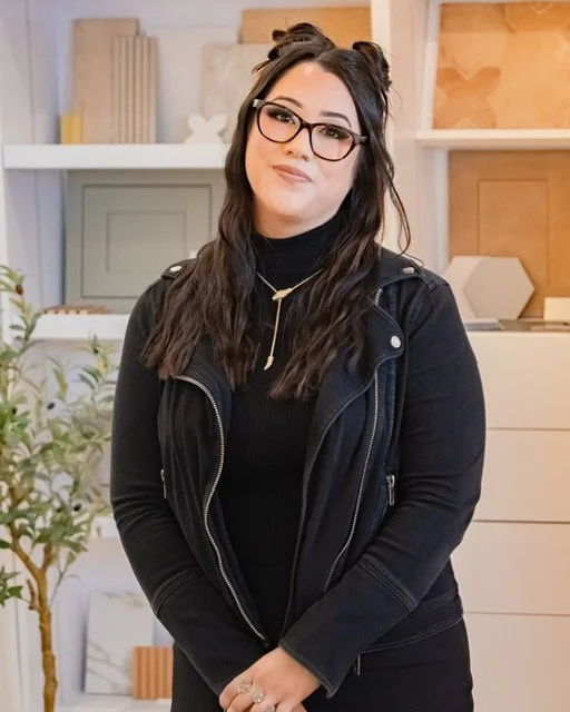 A woman with long dark hair and glasses posing indoors. She is wearing a black jacket and a black top, with a gold necklace. The background includes shelves with decorative items and a computer monitor.