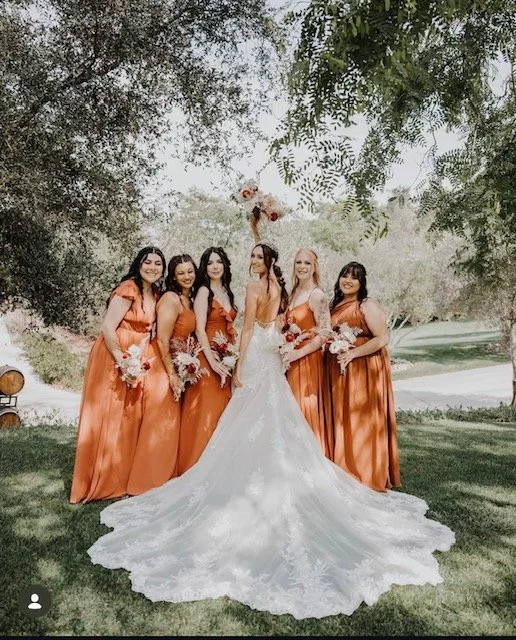 A bride in a white wedding dress standing with six bridesmaids in matching orange dresses outdoors, all holding bouquets, under a tree.
