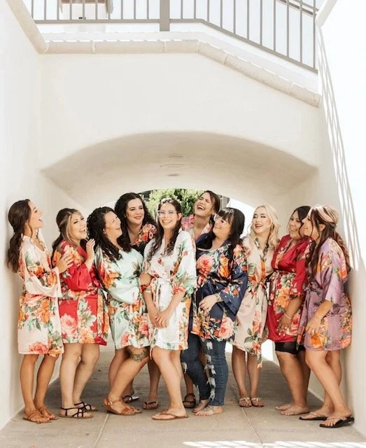 Group of women wearing colorful floral robes, standing outdoors under a white archway, smiling and laughing.