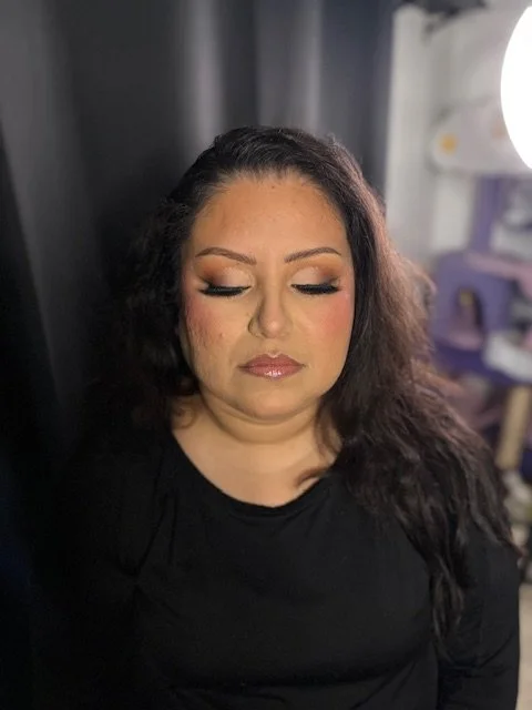 A woman with dark, wavy hair and makeup, wearing a black shirt, with closed eyes and a neutral expression, standing indoors against a dark background.