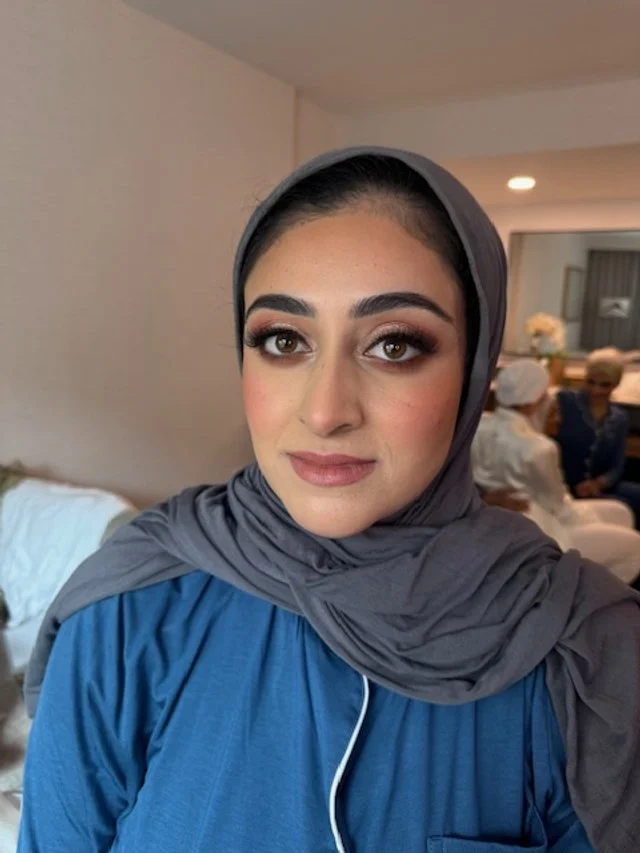 A young woman wearing a gray headscarf and a blue top, smiling softly at the camera, in a casual indoor setting with other people in the background.