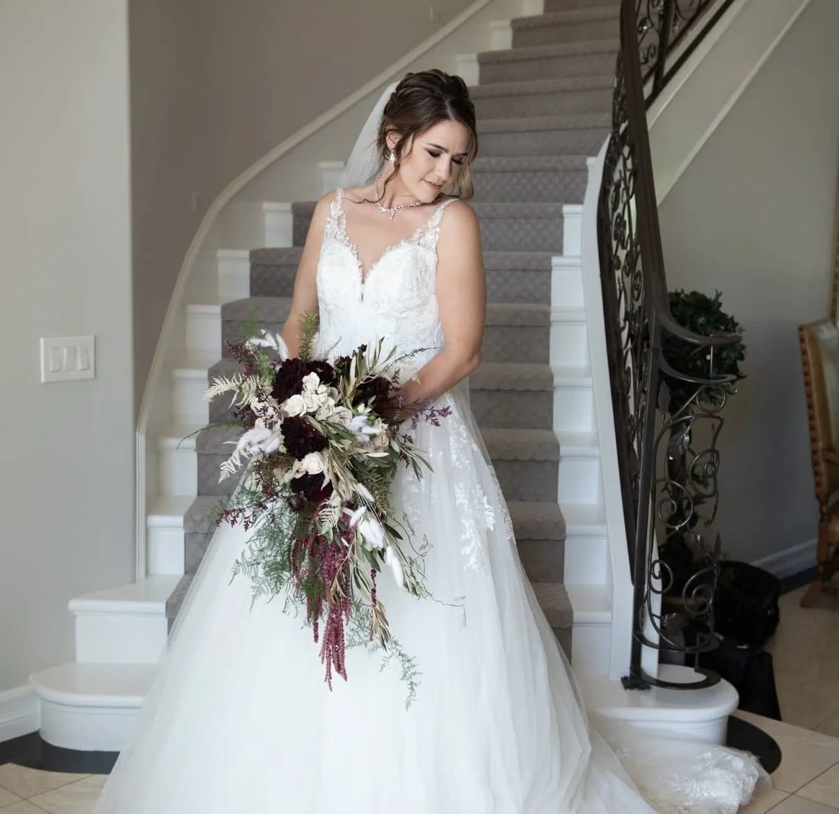 A bride in a white wedding gown standing on a staircase, holding a large bouquet of dark red and white flowers, with a neutral-colored interior background.