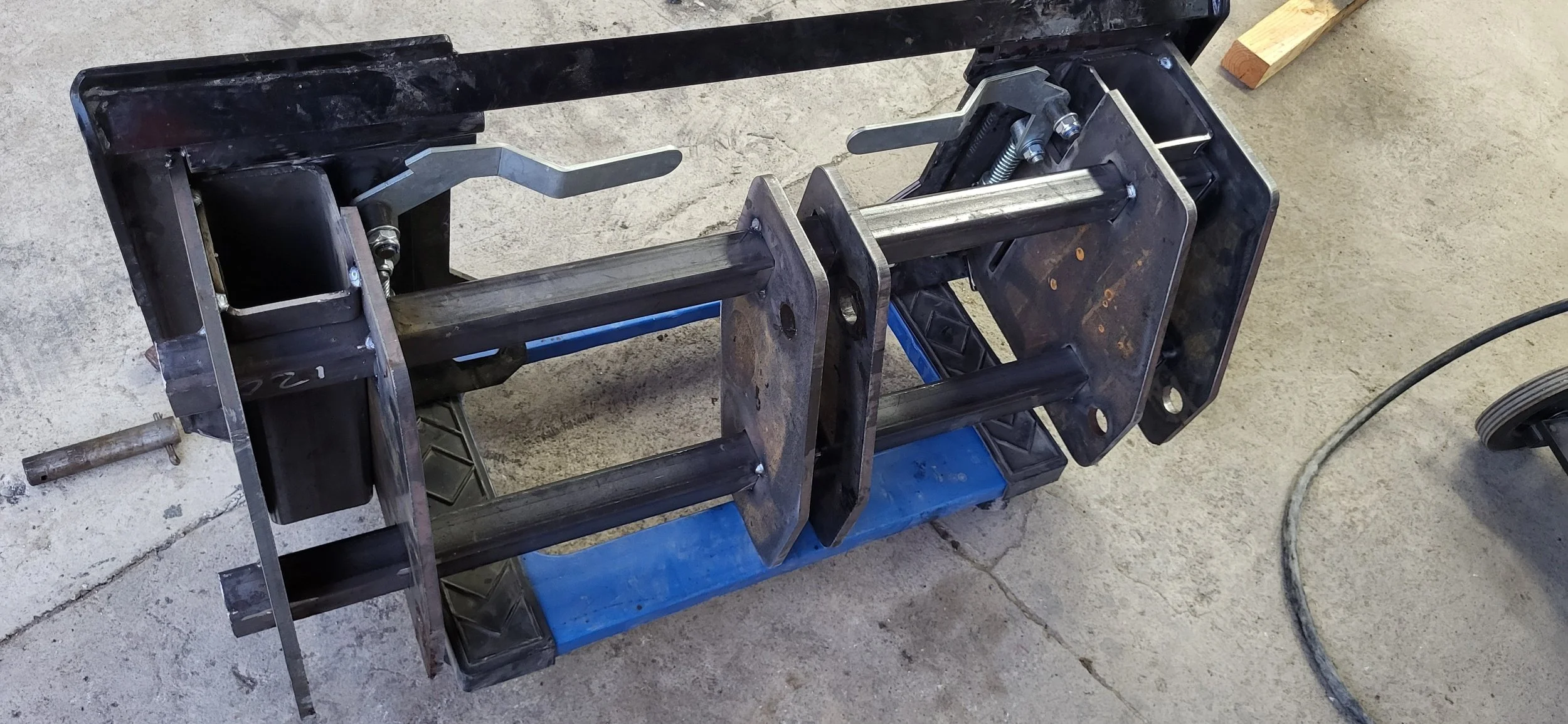 Metalworking vise with adjustable jaws mounted on a metal frame, with metal rods and screws, on a blue workbench in a workshop.