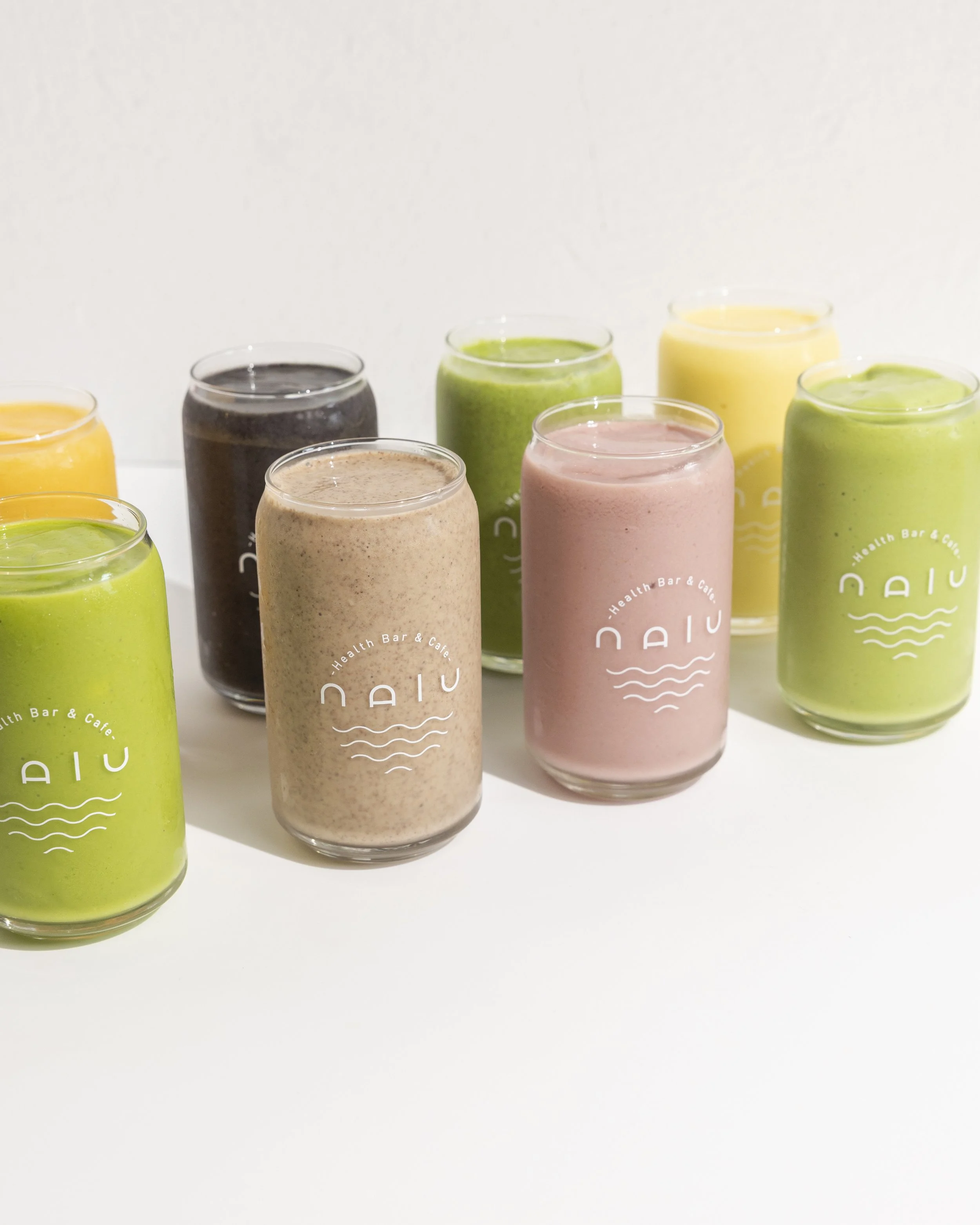 A yellow smoothie in a clear plastic cup with a black logo that reads 'Health Bar & Café'. In the background, there are a pineapple, an apple, and a coconut.