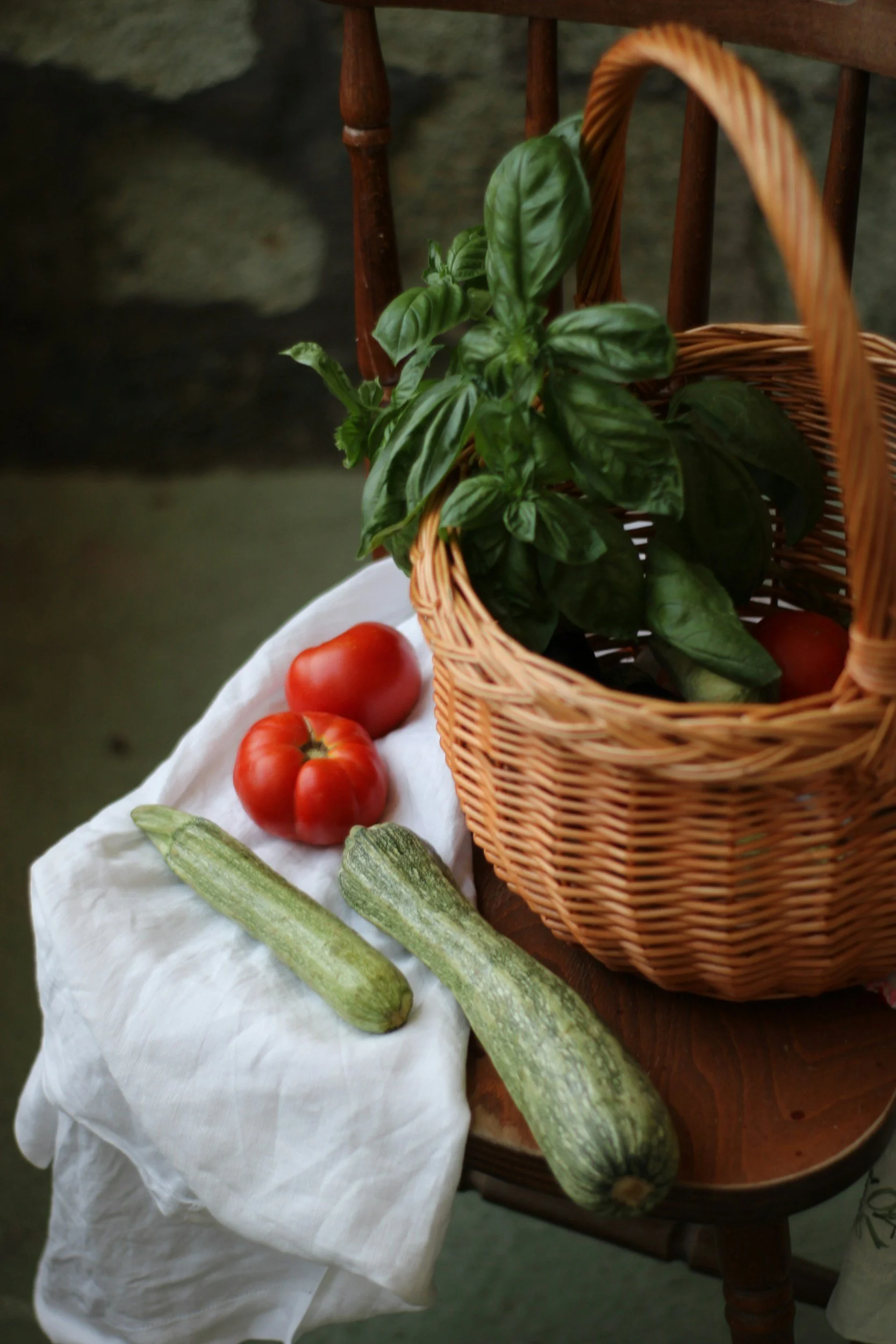 Fresh tomatoes, two zucchinis, and basil leaves on a white cloth and a wooden chair with a wicker basket containing more basil