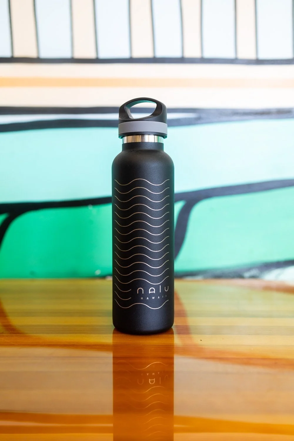 A black reusable water bottle with wavy white lines and the word 'NAIU' along with 'HAWAII' printed on it, placed on a wooden surface.