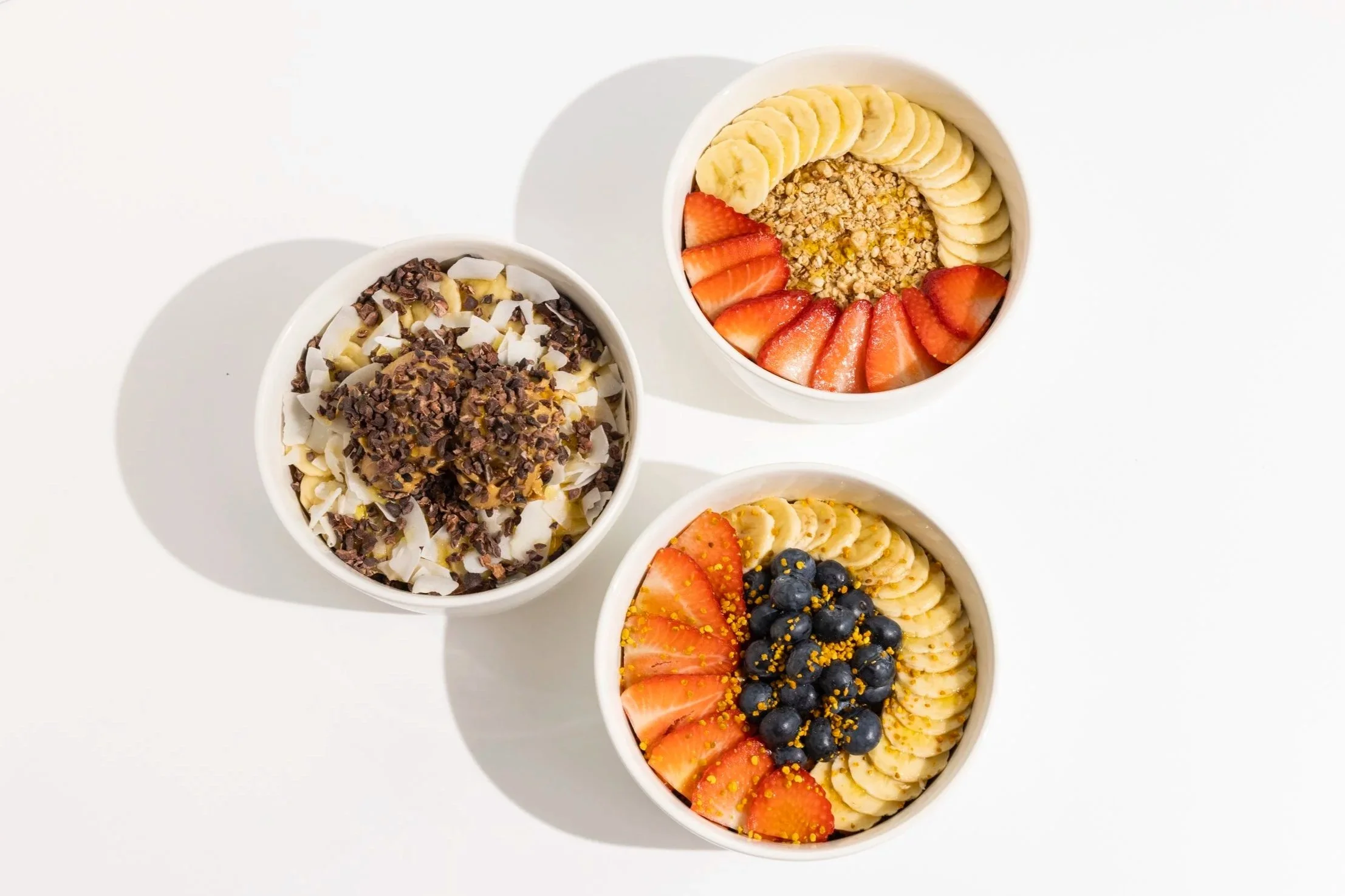 A cup of smoothie or acai bowl topped with sliced strawberries, blueberries, bananas, and sprinkled with granola on a green leaf, with additional fruit slices and blueberries on the side.