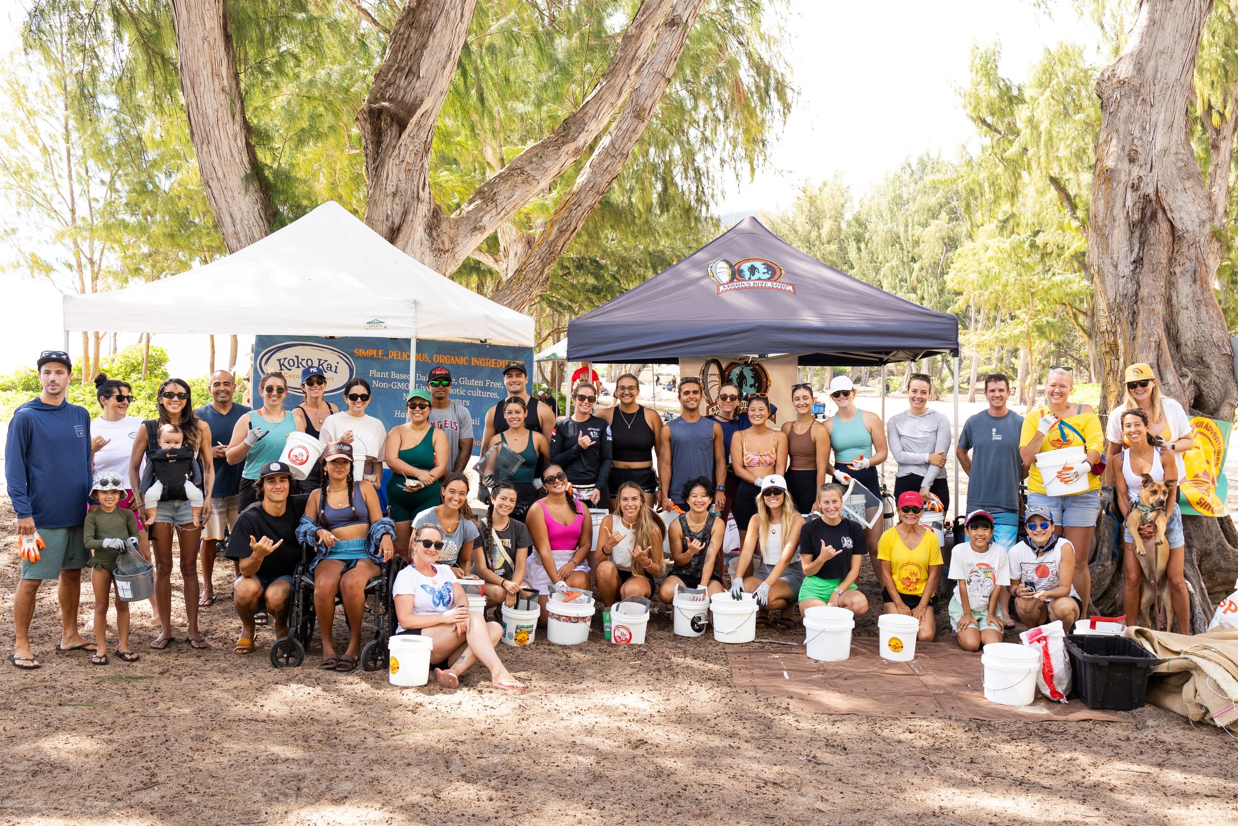A large group of people gathered outdoors under trees, posing for a group photo at an event with tents and banners. There are children, adults, and a dog, with some holding buckets or wearing hats and sunglasses. The setting appears to be a park or beach area.