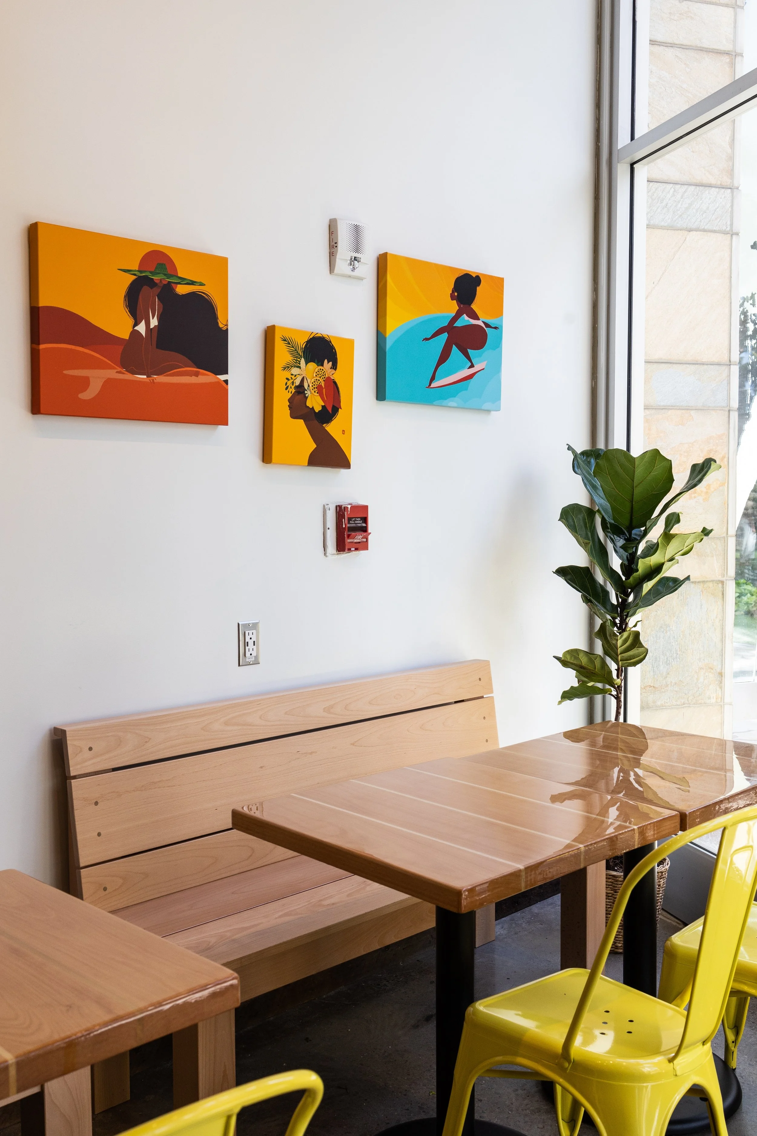 Interior view of a café with wooden tables, yellow chairs, a potted plant, and four colorful art prints of women on the white wall.